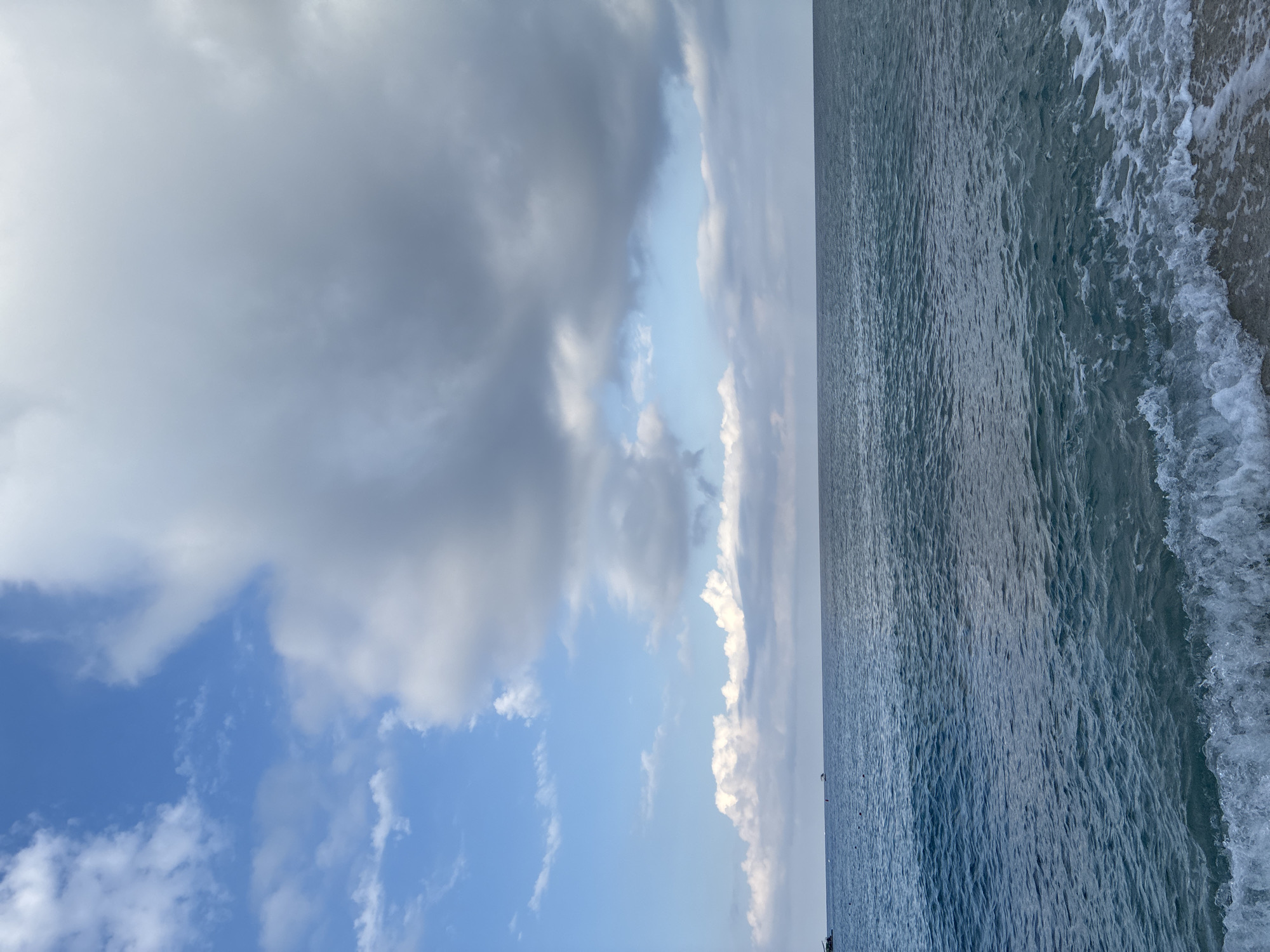 This image captures a scenic view of the Mediterranean Sea from the shore of Alanya, Turkey. The sky above is a blend of clear blue and dramatic, billowy gray clouds, suggesting dynamic weather conditions. The sea itself is a deep, shimmering blue, with small waves lapping at a sandy beach in the foreground. In the distance, the horizon is calm, with a faint line indicating where the sea meets the sky. There are small boats visible on the water, and a few buoys dot the surface closer to shore, suggesting this is an area used for water activities or navigation. The overall impression is one of natural beauty and tranquility, characteristic of the coastal landscapes found in Alanya.