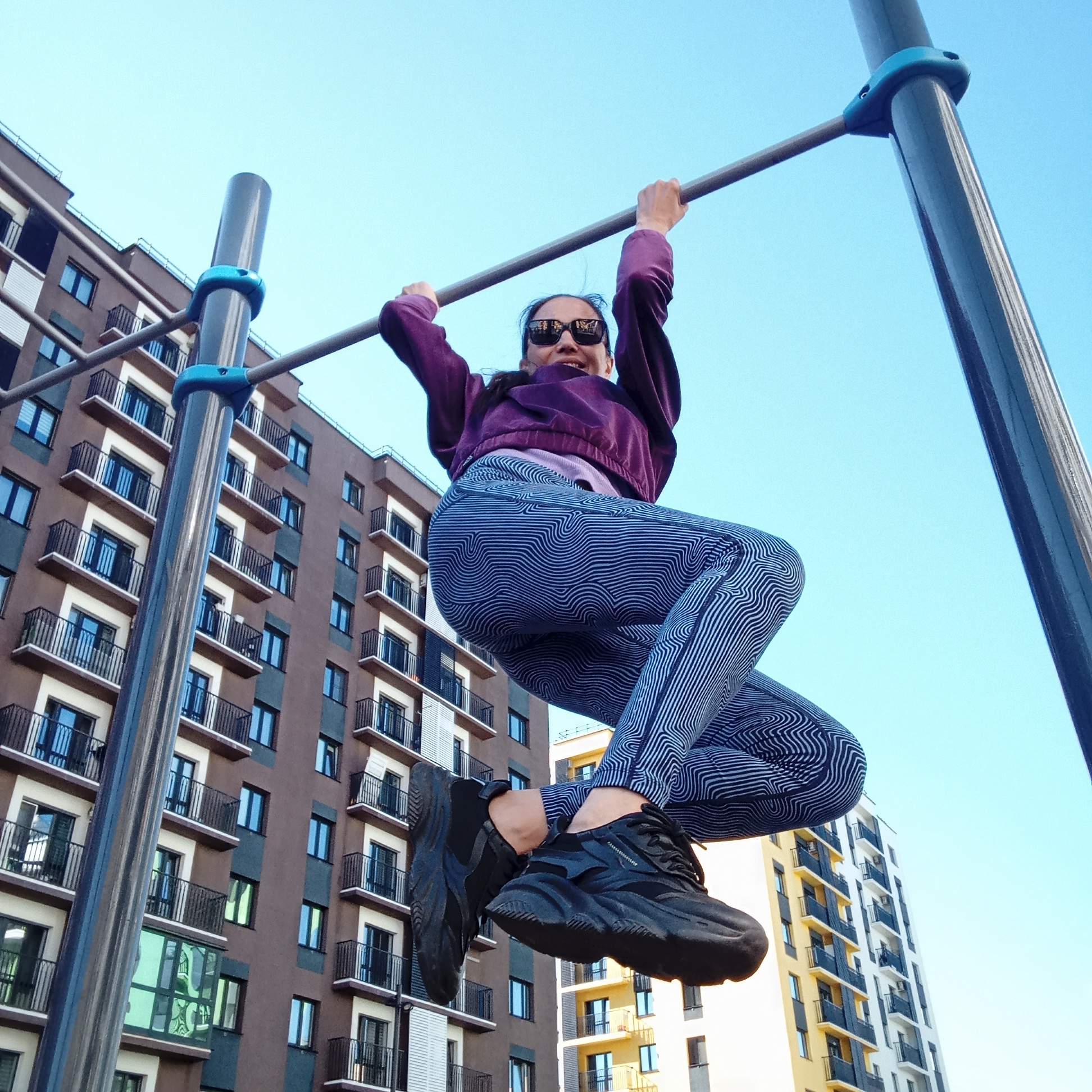 A low-angle outdoor shot captures a person actively hanging from a horizontal bar. The individual is wearing dark sunglasses, a purple long-sleeved top, and distinctive leggings with a blue and white wavy line pattern. They are also wearing chunky black athletic shoes. Their dark hair is pulled back, and their mouth is open in what appears to be a smile or laugh, suggesting enjoyment or playful exertion. They grip the grey metal bar with both hands, with their legs bent and pulled upwards.
The workout structure features grey horizontal bars connected by bright blue clamps to vertical posts. In the background, under a clear, bright blue sky, stand multiple modern residential buildings. A prominent multi-story building to the left has a brown facade with cream-colored sections surrounding numerous balconies, which are fitted with black railings. Another building, partially visible to the right and further back, appears to be yellow and white. The bright conditions indicate it is daytime. The scene depicts an active moment in an urban residential or park setting. No visible text is present in the image.