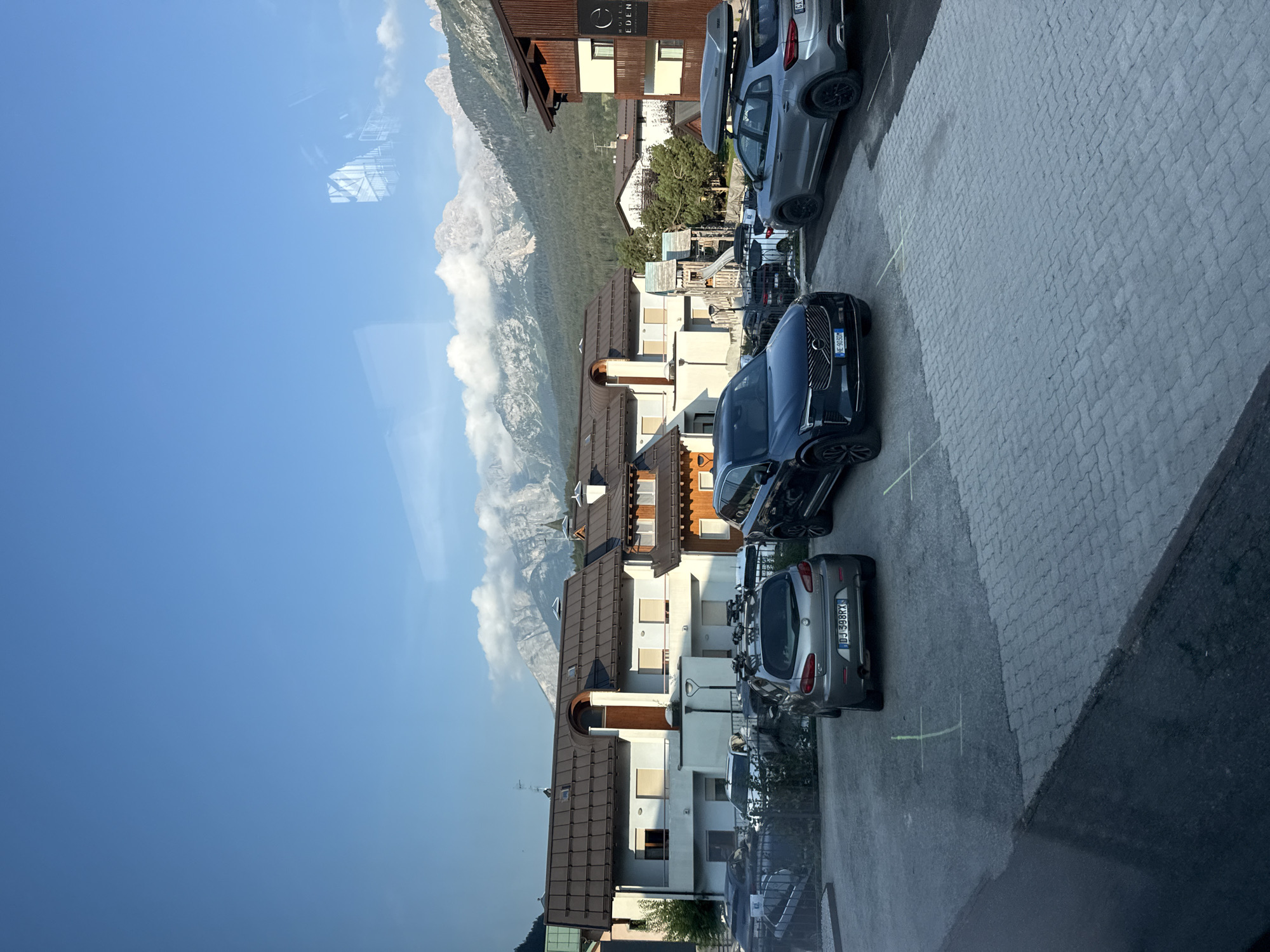 The image shows a parking lot in front of buildings with mountains in the background. Several cars are parked in the lot, including a dark blue SUV, a silver hatchback with a roof rack, and a silver SUV with a cargo box on its roof. The buildings appear to be residential or hotels, with balconies and windows. The mountains in the background are rocky and covered in trees at the base, with some clouds near the peaks. The sky is blue and clear. The scene is set in Andalo, Italy, as indicated by the presence of the Hotel Eden sign on one of the buildings. There are no people visible in the image. The parking lot is paved with interlocking stones.