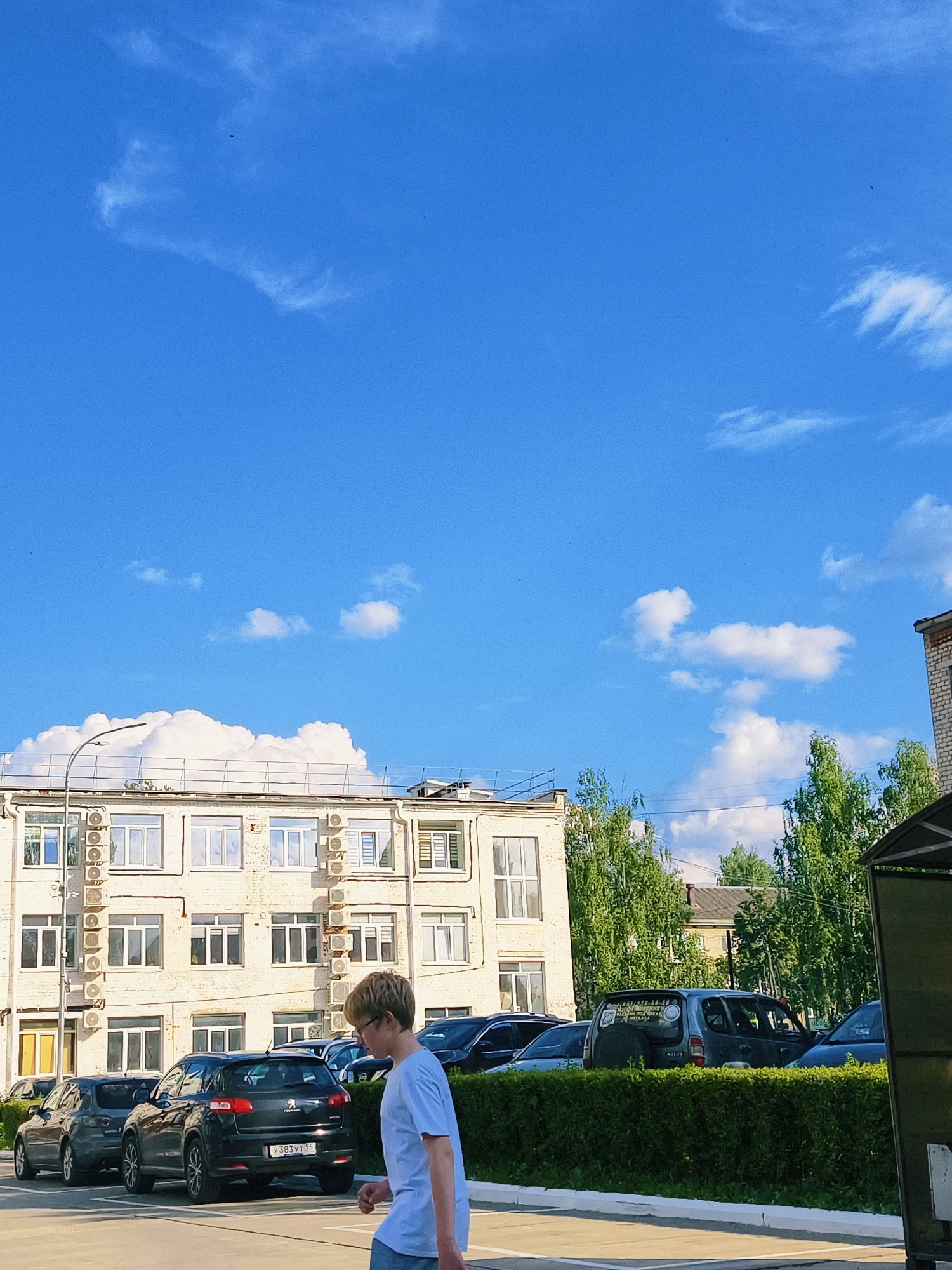 In Chaikovsky, Russia, on a bright sunny day with a clear blue sky dotted with sparse white clouds, a young boy with blond hair and glasses walks from right to left in the foreground. He is wearing a light blue t-shirt and gray shorts. Behind him, a row of parked cars is visible, including a black Peugeot SUV in the center with a Russian license plate that reads "Y 383 VT 96". To the left of the Peugeot, a dark gray car is parked, and further left, another dark gray car. To the right of the Peugeot, another dark car is partially visible.
In the background, a light-colored, multi-story building stands. The building has numerous windows, and air conditioning units are mounted below some of them. A tall street lamp is positioned to the left of the building. Trees with lush green foliage line the area behind the cars and the building. A partially obscured structure, possibly a bus stop shelter with a green roof, is visible on the far right of the frame. The scene appears to be a parking lot in an urban or semi-urban environment.