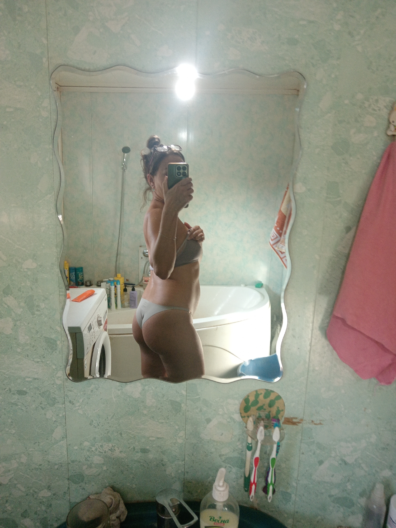 A woman is taking a selfie in a bathroom mirror. She is wearing a grey bra and grey underwear and has her hair up in a bun. She is standing next to a bathtub and a washing machine. The bathroom walls are tiled with a light green marble pattern. A pink towel hangs on the wall to the right of the mirror. There are toothbrushes and a bottle of soap on the counter below the mirror. The overall impression is of a residential bathroom in a modest dwelling.