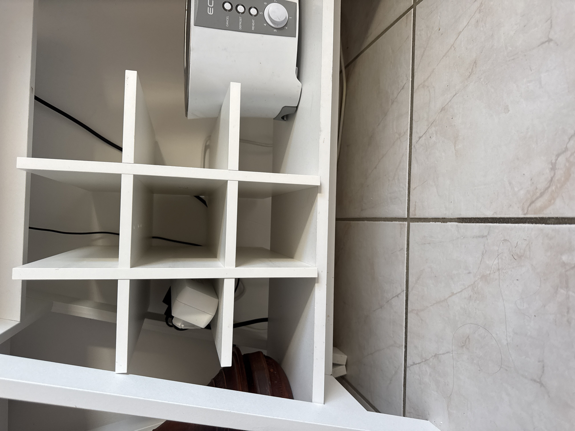 The image depicts the interior of a white shelving unit, likely a media console or a storage unit. The unit has multiple compartments. In the top left compartment, a white power adapter with a cord is visible. To its right, in the top middle compartment, a black cable is draped. The second shelf from the top, on the left, contains a dark brown decorative item. On the second shelf from the top, on the right, a white toaster with silver accents is placed. The shelf below the toaster is empty. The bottom shelf of the unit is also visible, with its front edge slightly visible. The unit itself is positioned on a floor covered with light-colored tiles, separated by dark grout lines. Some dark strands, possibly hair or dust, are scattered on the floor in front of the shelving unit. The scene is indoors, and there is no indication of the city of Höhenkirchen-Siegertsbrunn, Germany within the image itself.