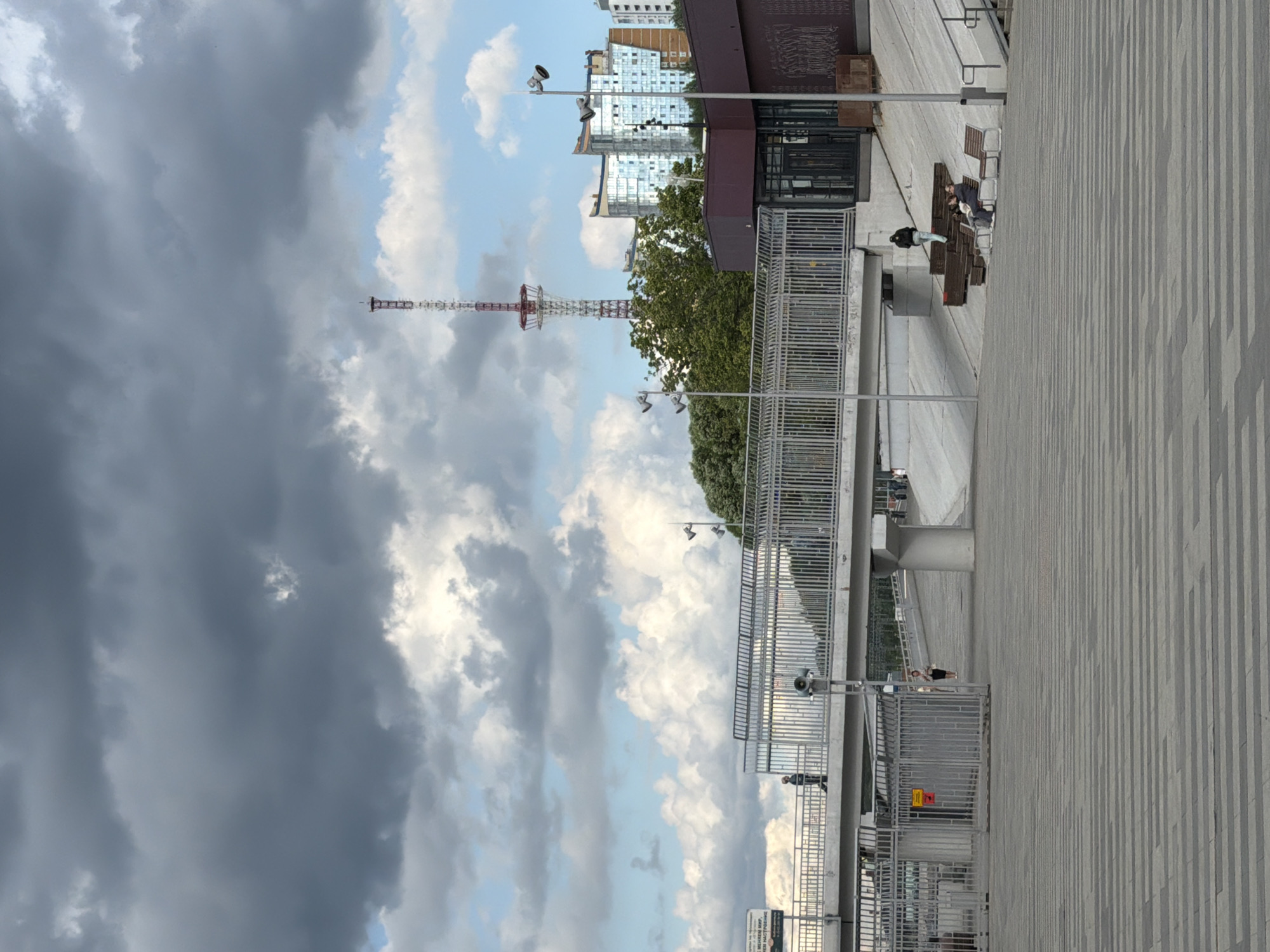 This media file depicts an outdoor scene with a mix of urban and natural elements. The foreground is dominated by a patterned concrete walkway, possibly part of a plaza or promenade, that extends diagonally across the frame. Several metal benches are visible along the edge of this walkway, with some people seated and others standing nearby.
Further back, a prominent feature is a tall, lattice-like structure that appears to be a telecommunications tower, painted in red and white sections. It is set against a sky filled with dramatic clouds, ranging from dark, brooding storm clouds on the left to brighter, sunlit clouds on the right.
Behind the tower and scattered buildings, a lush green tree adds a natural element to the urban landscape. The buildings in the background are a mix of modern high-rises with glass facades and older, more industrial-looking structures.
The overall impression is one of a public space, possibly a waterfront or a park adjacent to a city. The weather appears to be changeable, with both dark clouds and patches of blue sky and sunlight suggesting a dynamic day. The presence of people suggests that the area is in use, likely during daylight hours. The context provided places this scene in Perm, Russia, which helps in understanding the architectural styles and urban environment. No specific event is clearly indicated, but the scene suggests a typical day in a city.
