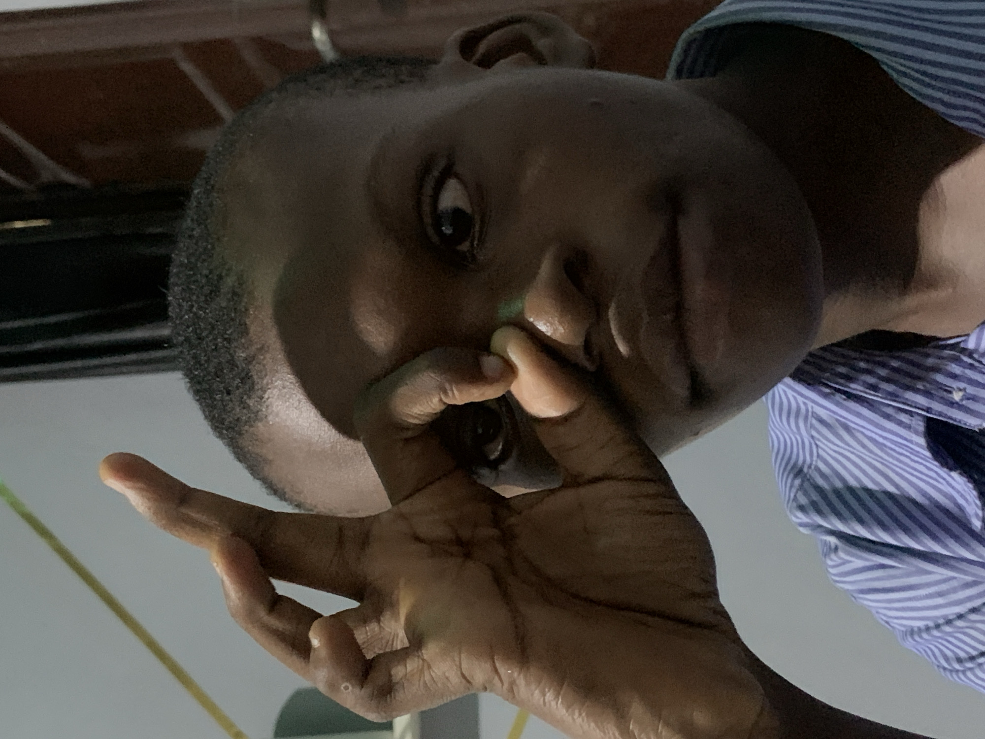 A close-up shot shows a person of African descent, wearing a blue and white striped shirt, holding their hand up to their face. The person's eyes, nose, and hand are prominent. The background is blurry. The photograph's location is provided as Ibasa, Nigeria, but no element of the image allows to confirm the city.