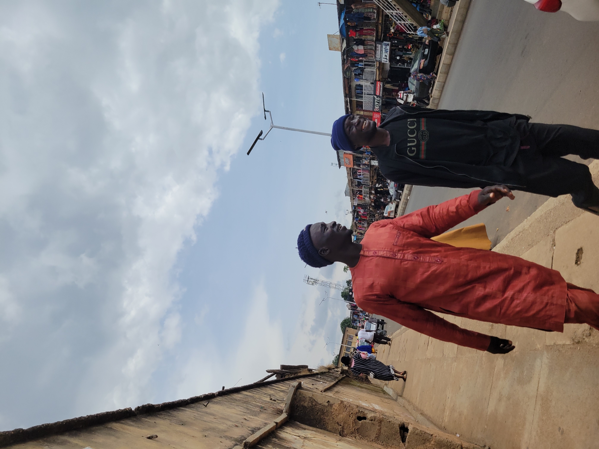 Two young men walk side-by-side on a concrete sidewalk next to a busy street. The man on the left wears a red traditional African garment with a round neckline and buttons down the front. He also wears a blue beanie and black gloves. The man on the right wears a black tracksuit with a blue beanie. A yellow cloth is visible between the two men, partially obscured by the man in red. In the background, there is a bustling marketplace with stalls selling clothing, and cars driving on the street. The sky is overcast with scattered clouds.