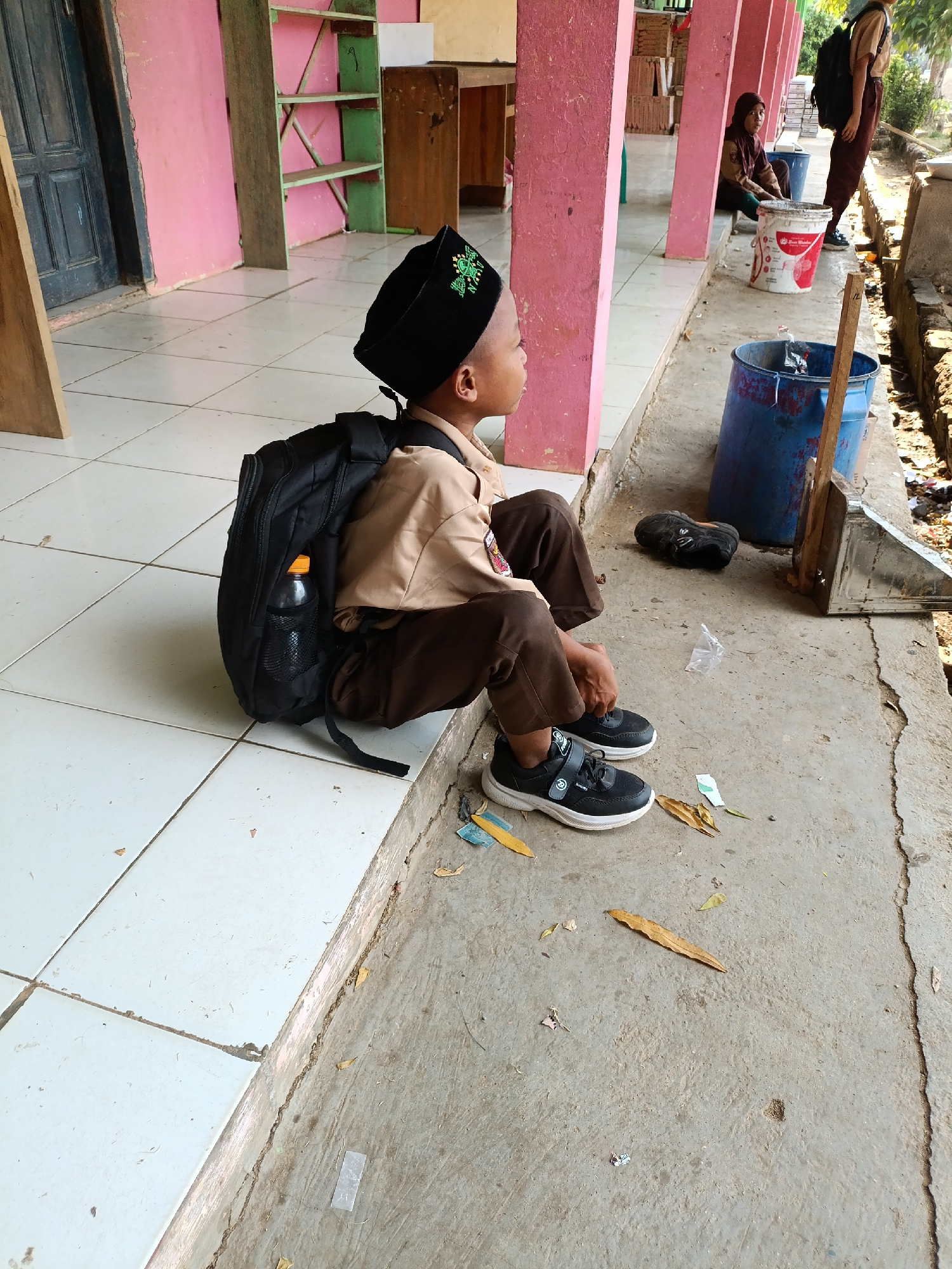 A young boy in a brown uniform and a black skullcap with green embroidery sits on a tiled floor next to a concrete pathway. He is wearing black sneakers and has a black backpack with a water bottle inside on his left side. The boy is facing right, looking away from the camera. Behind him, a pink wall with support beams is visible, leading to a hallway with shelves and doors. Further down the hallway, another person in a similar uniform is seated, and two large blue and white buckets are present. The ground is littered with leaves and debris. This scene appears to be in a school environment in Sumberejo, Indonesia.