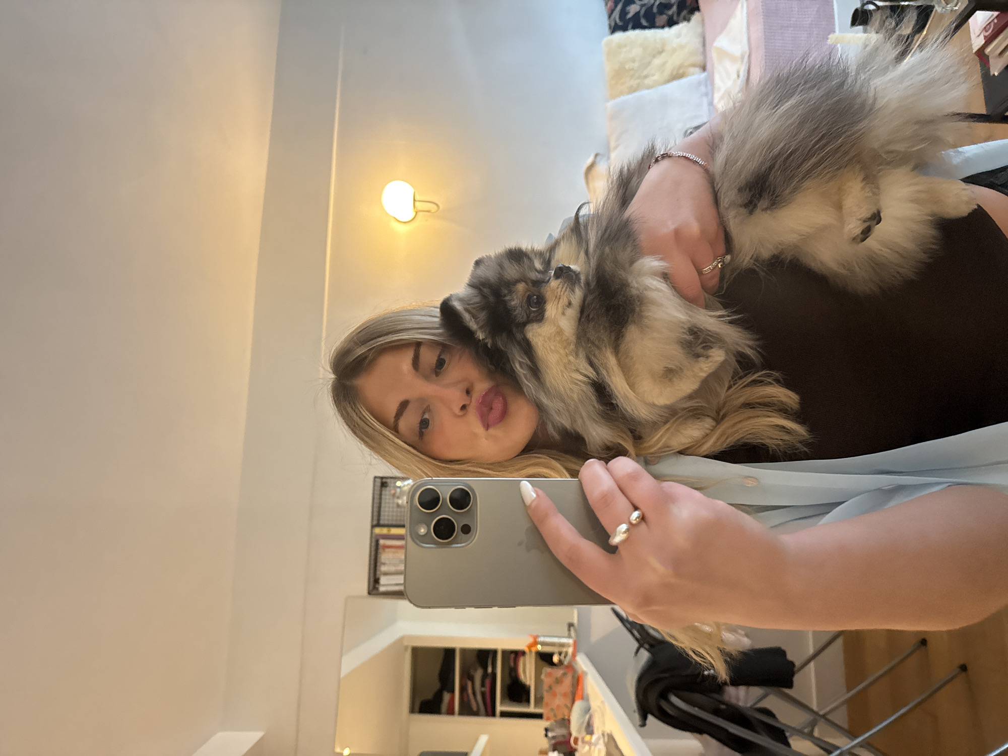 This mirror selfie captures a young woman holding a small, fluffy dog indoors. The woman, with long blonde hair, appears on the right, looking directly at the camera with a playful "duck face" expression. She is wearing a light-colored top over a darker garment. In her left arm, she cradles a grey, black, and white Pomeranian-like dog, whose head is resting peacefully near her face. The dog appears calm and comfortable.
The woman holds a grey iPhone, identifiable by its three camera lenses, in her right hand, positioned in the lower-middle part of the frame. She sports a distinctive silver ring on her right ring finger and a rose-gold colored bracelet on her left wrist. Her fingernails are painted white.
The setting appears to be a cozy indoor space, possibly a home in Paris, France. A warm, illuminated wall-mounted light fixture casts a soft glow on the upper left side of the background, suggesting artificial lighting or evening hours. Behind the woman, glimpses of furniture, possibly a bed or sofa adorned with cushions, are visible. The mirror's reflection below the phone reveals shelves or storage units with various items, indicating a lived-in domestic environment.