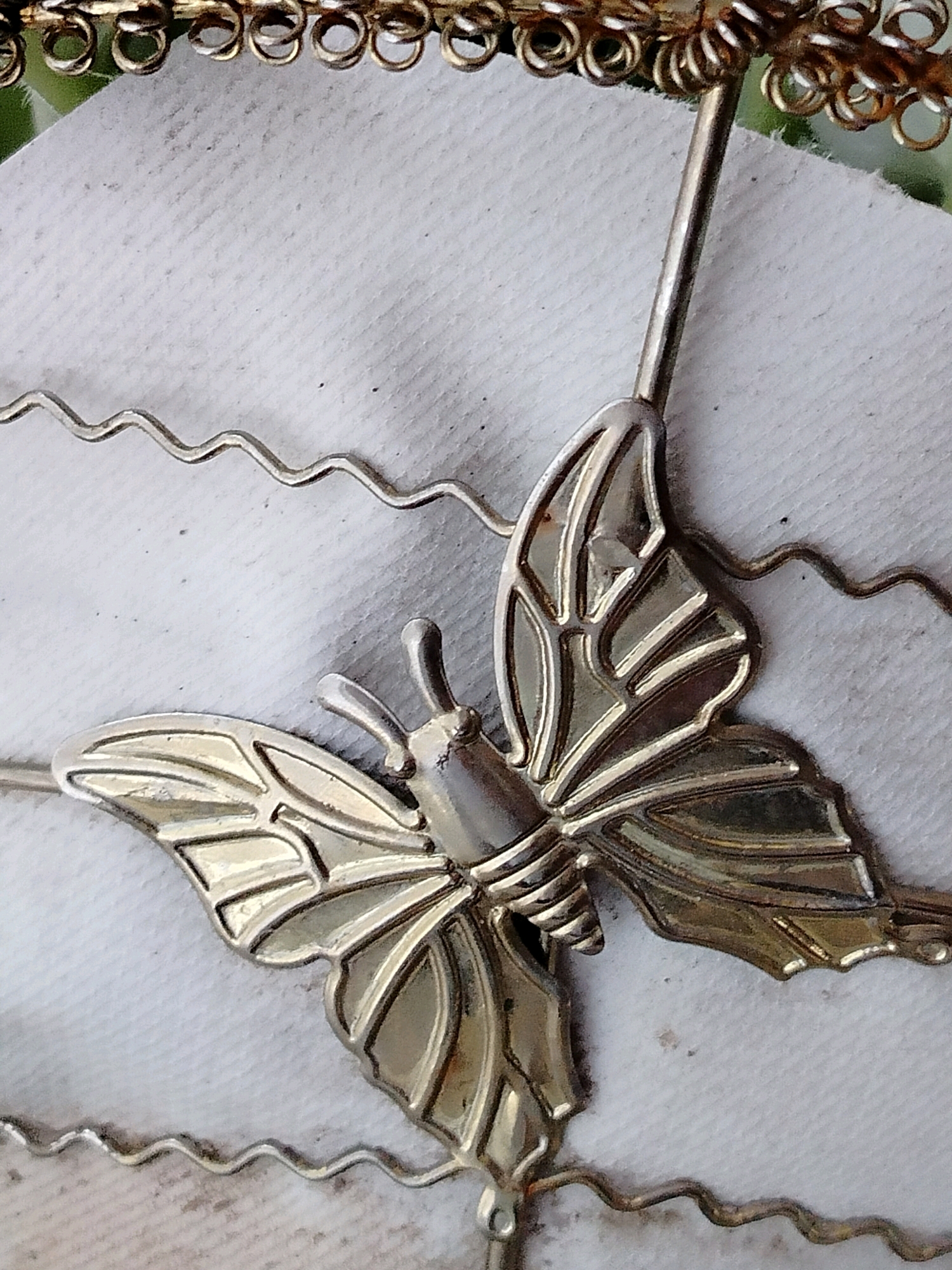 A close-up, overhead shot shows a metallic, silver butterfly ornament resting on a white, textured surface, possibly fabric or paper. The butterfly is detailed with etched lines on its wings, simulating veins. A thin, silver wire, possibly part of a larger decorative piece, extends upwards from the butterfly's body and curves out of frame at the top. Below the butterfly, a thin, wavy silver wire also runs horizontally across the frame. The background is entirely out of focus, but appears to be a neutral, light color with subtle shadows and possibly some faint green elements at the very top, suggesting it might be part of a larger item or setting. There are no people, actions, or interactions depicted. The image is taken indoors, with the lighting suggesting a diffused light source. The location in the city of Көлкент, Kazakhstan, is not visually indicated in the image.