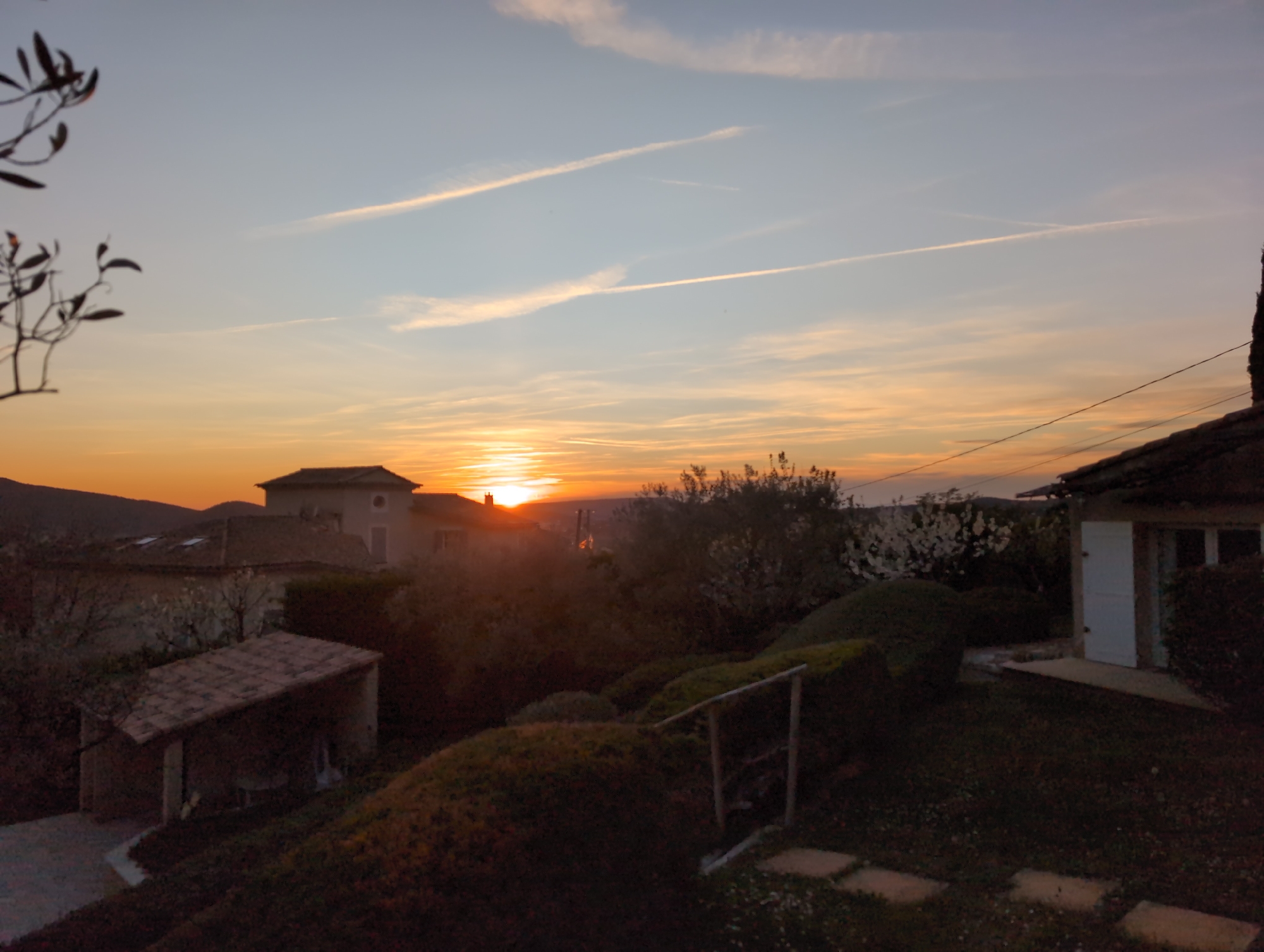 The image depicts a sunset in Saint-Marcellin-lès-Vaison, France. The sun is setting behind hills in the distance, creating an orange glow in the sky. Above the sunset, there are several contrails. In the foreground are buildings with tiled roofs, trees, shrubs, and a garden area with stepping stones. A small, open structure with a tiled roof is visible to the left, and a building with a white door and window is visible to the right. A few branches of a tree are visible in the upper left corner.