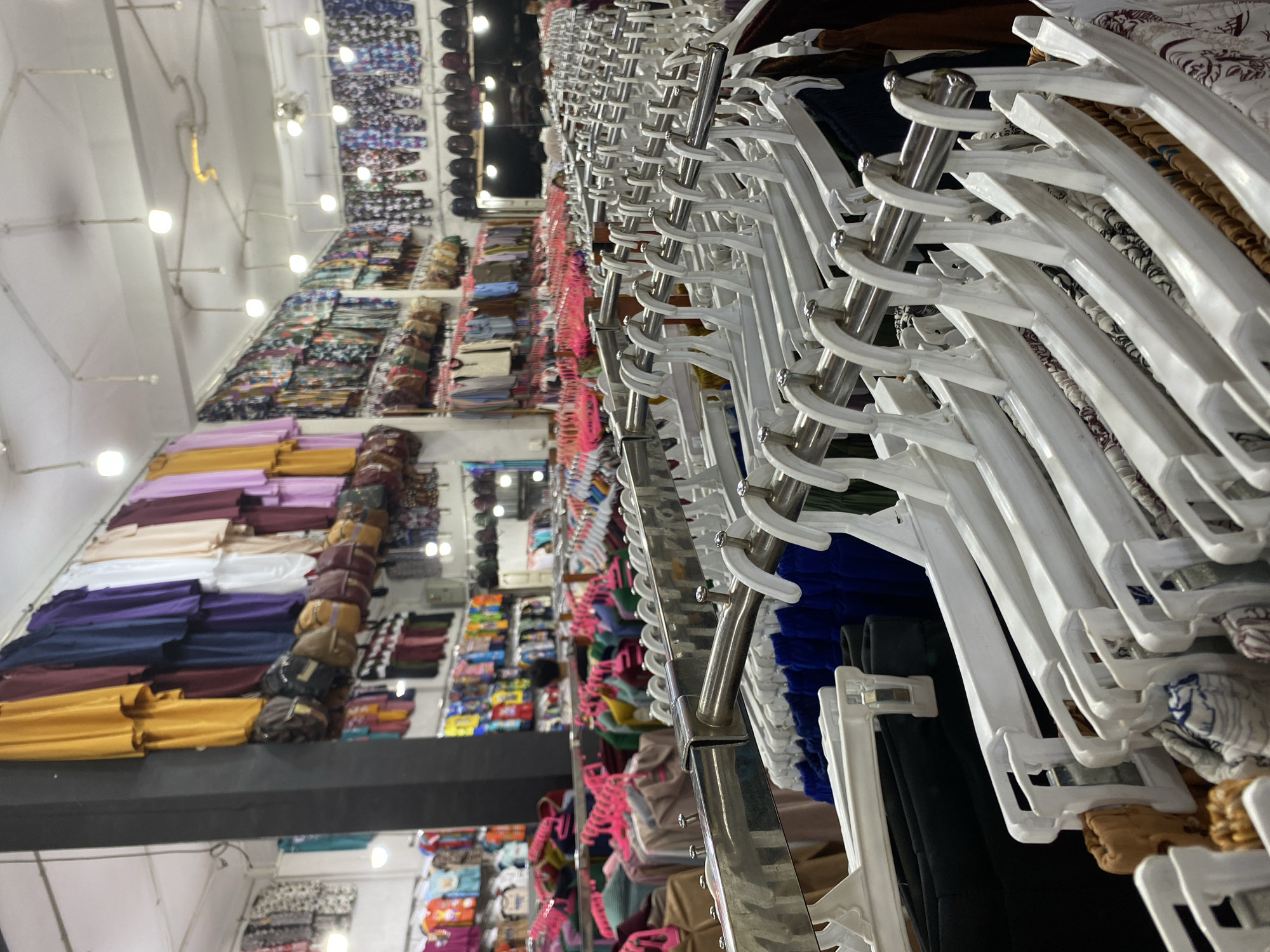 This is a medium-shot, eye-level, indoor photograph of a clothing store, likely a boutique or a stall, filled with various garments. The store is brightly lit by fluorescent lights on the ceiling. The foreground is dominated by rows of clothing hanging on white plastic hangers suspended from metal racks. The garments are predominantly trousers and skirts, displaying a variety of patterns, including floral and geometric designs in colors such as blue, brown, white, and black. Some items appear to be folded on shelves or racks further back in the store.
In the background, more clothing is visible, with several rows of garments hanging on pink hangers, suggesting a section for tops or dresses. These garments are in solid colors like yellow, purple, navy blue, and white, as well as printed fabrics. Bags are also displayed hanging above some of the clothing racks, adding to the retail environment. The walls of the store are lined with more clothing displays and shelving. The overall impression is one of a well-stocked and organized retail space. The location, Lamteumen Barat, Indonesia, implies a tropical or subtropical climate, which might influence the types of clothing available, such as lighter fabrics and styles.