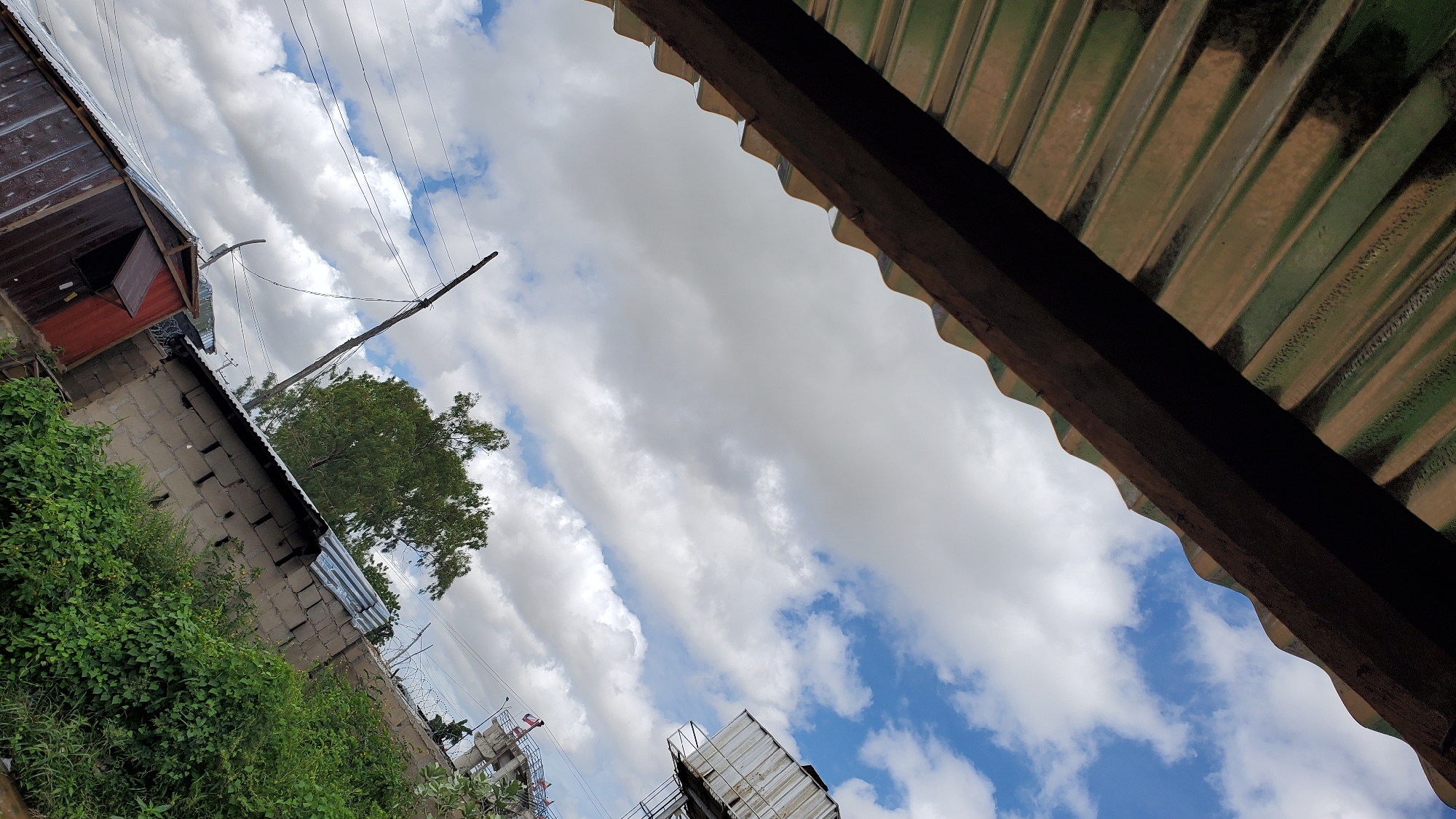 The image depicts a view looking upwards from beneath a corrugated metal roof overhang, revealing a cloudy sky. In the foreground, the textured green and brown corrugated metal roof with a dark wooden beam is prominent, angled diagonally across the frame. Below this, the scene opens to a wider view of the sky, dotted with white clouds against a blue backdrop.
Further into the scene, a variety of structures and natural elements are visible. To the left, a smaller, dark red metal-clad structure with an open window or vent is partially visible. Adjacent to this is a wall constructed from grey cinder blocks, topped with barbed wire. Dense green vegetation, likely bushes and trees, grows along this wall and in front of it.
In the mid-ground and background, more of the cloudy sky is present, with a large, leafy green tree rising towards it. A tall, thin wooden pole with wires extending from it stands vertically in the mid-ground. In the upper right portion of the image, a large, rectangular metal tank or structure is perched atop a reinforced concrete support, suggestive of water infrastructure or a similar utility. Further in the distance, another concrete structure, possibly a tower or bridge support, is visible with small flags.
The scene appears to be an outdoor area in a semi-urban or rural setting, likely within Jalingo, Nigeria, given the architectural style of the buildings, the vegetation, and the sky conditions typical of the region. There are no people or discernible actions taking place. The overall impression is one of an everyday scene viewed from a sheltered vantage point.