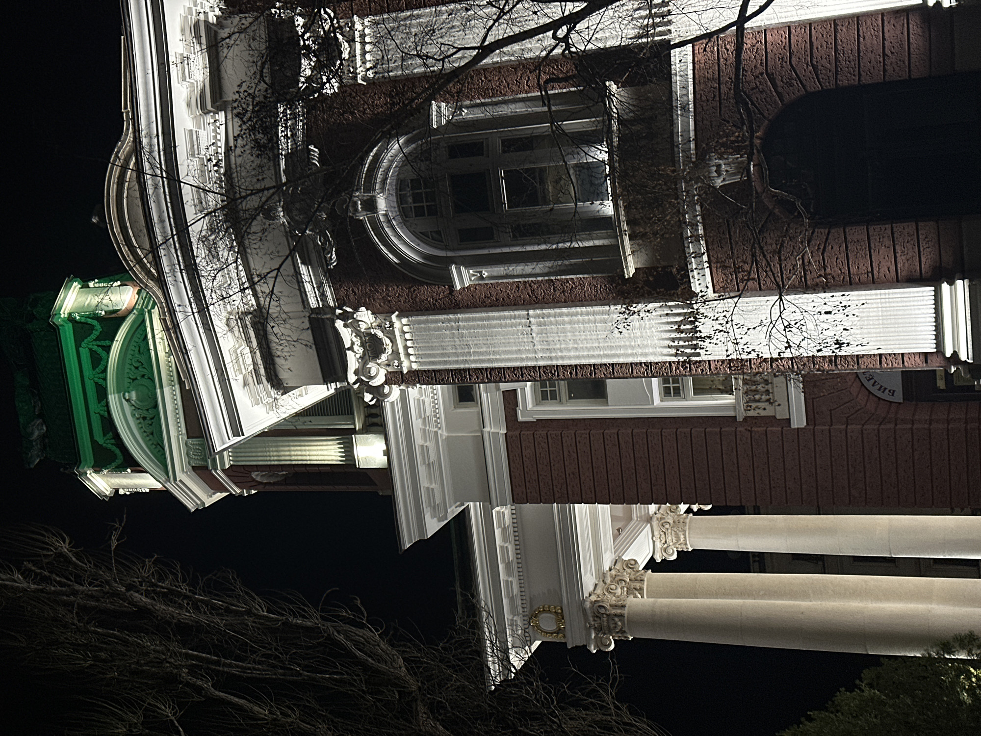 The image shows a building at night in Sofia, Bulgaria. The building has classical architectural elements, including columns, ornate window frames, and decorative molding. A green-lit section is visible on the left side of the building. Bare tree branches are partially obscuring the building.