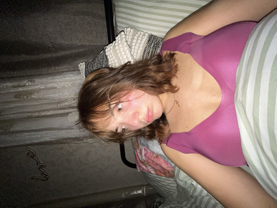A young woman reclines in bed, gazing downward with a slightly melancholic expression. She has auburn hair, lightly tanned skin, and wears a simple gold necklace with a cross. The woman is partially covered by striped bedding and a pink top is visible. The background includes a textured headboard, and a "love" wall decoration is noticeable in the lower left corner. The overall lighting is soft and dimly lit.