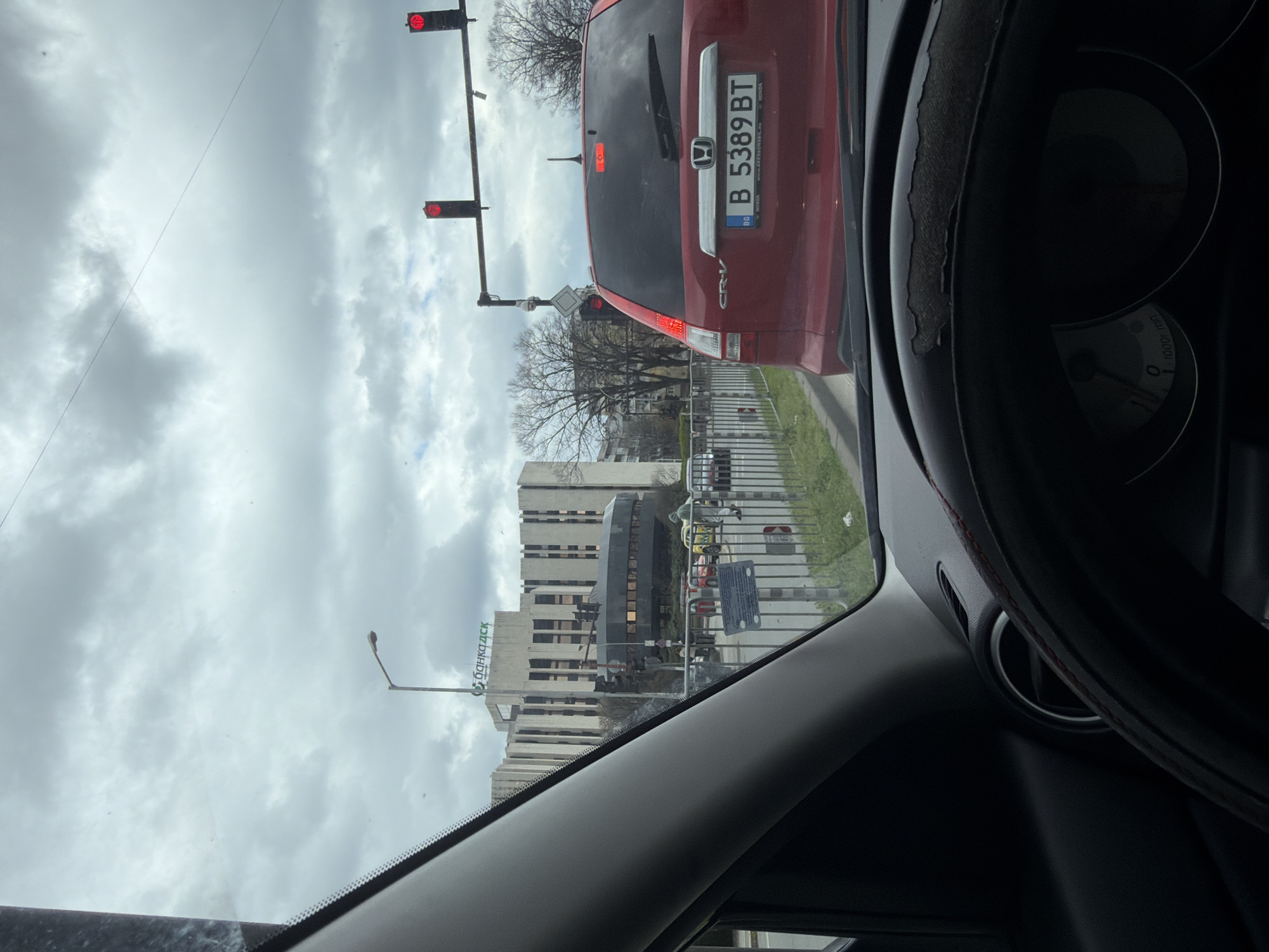 The image, taken from inside a car, shows a view through the windshield at a traffic light that is red. In front is a red Honda CRV with the license plate "B 5389 BT". The building in the background has a sign "Банка ДСК." The speedometer reads 0. The location is likely in the city of Varna, Bulgaria, given the license plate and the Cyrillic script on the building. The environment appears overcast.