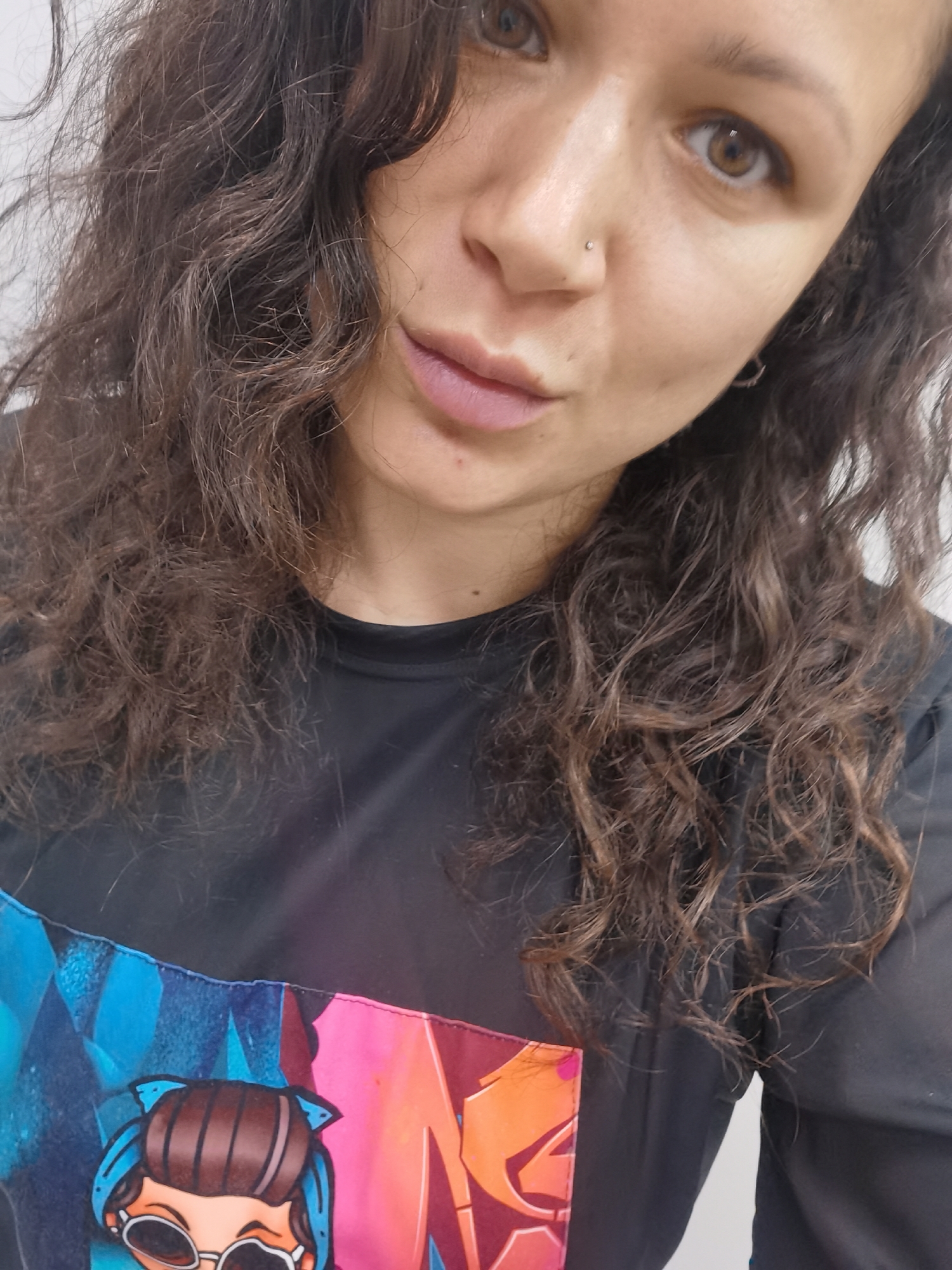 A woman with dark curly hair is pictured in a close-up selfie. She has light brown eyes and a nose piercing. She's wearing a black top with a colorful graphic on the front featuring a cartoon character with sunglasses. The background is a plain, light-colored wall. There is no indication in this image that the location is Chaikovsky, Russia.