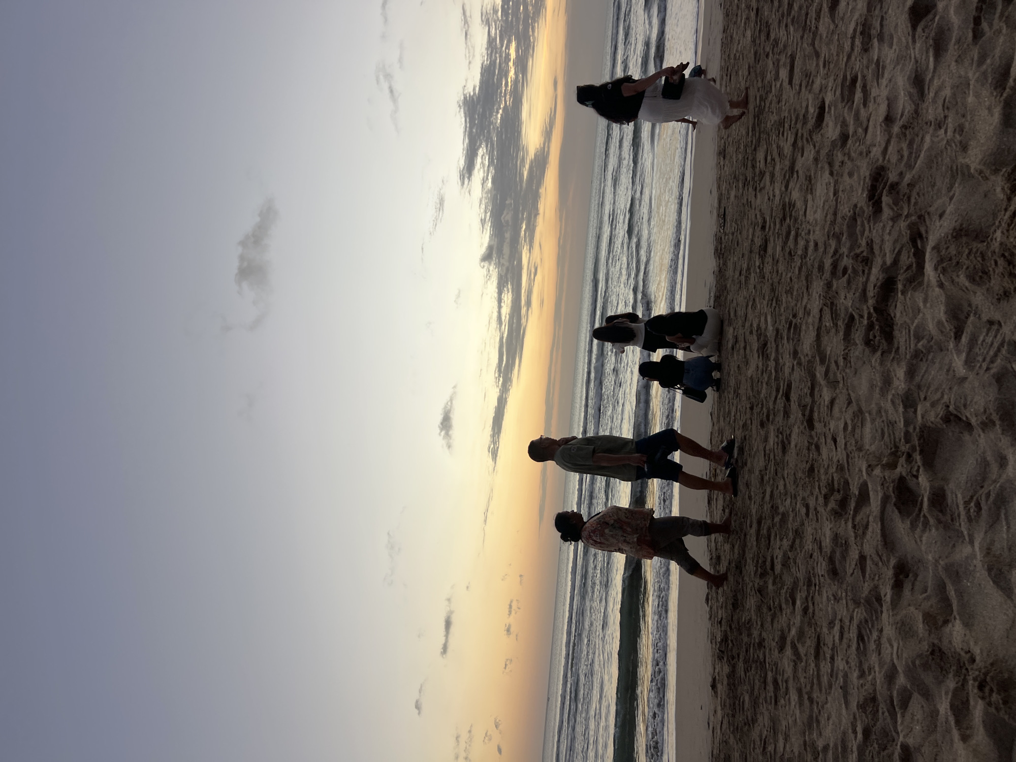 The image depicts a beach scene in Kuta, Indonesia, during what appears to be sunset or dawn. The sky is a gradient of soft blues, grays, and pale oranges, with scattered clouds. The ocean is visible in the background with gentle waves rolling onto the sandy shore. Several people are present on the beach.
On the left side of the image, a couple is walking. The man is wearing a green t-shirt and blue shorts, and the woman is wearing a patterned shirt and pants. Further to the right, three women are standing and looking out at the ocean. Two of them are wearing dark tops and light-colored bottoms, and one is wearing a white skirt and black top. One woman in the group is holding a black bag. In the foreground, a woman in a white skirt and black top is walking away from the camera towards the right. Another woman is partially visible in the far right, also walking away. Two individuals are seated on the sand in the middle ground, one wearing dark clothing and the other wearing a white outfit. The sand in the foreground is textured with footprints and ripples. The overall atmosphere is tranquil and serene, with the soft lighting characteristic of twilight.