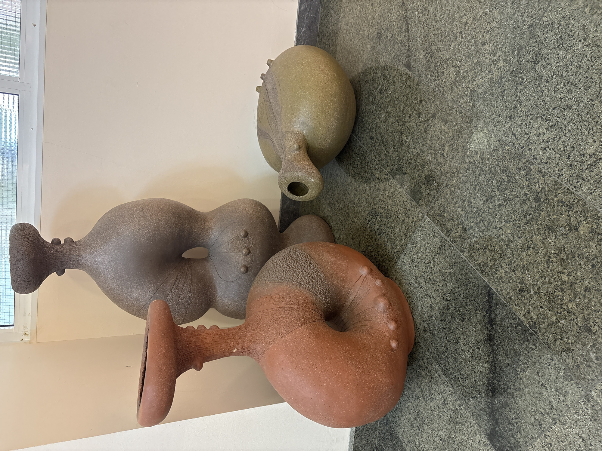 The image shows three irregularly shaped ceramic objects lying on a gray, speckled floor next to a light-colored wall with a window. The objects are different colors: one is light brown, one is gray, and one is reddish-brown. They all have bulbous bodies and elongated necks. The location is Unknown, Unknown.
