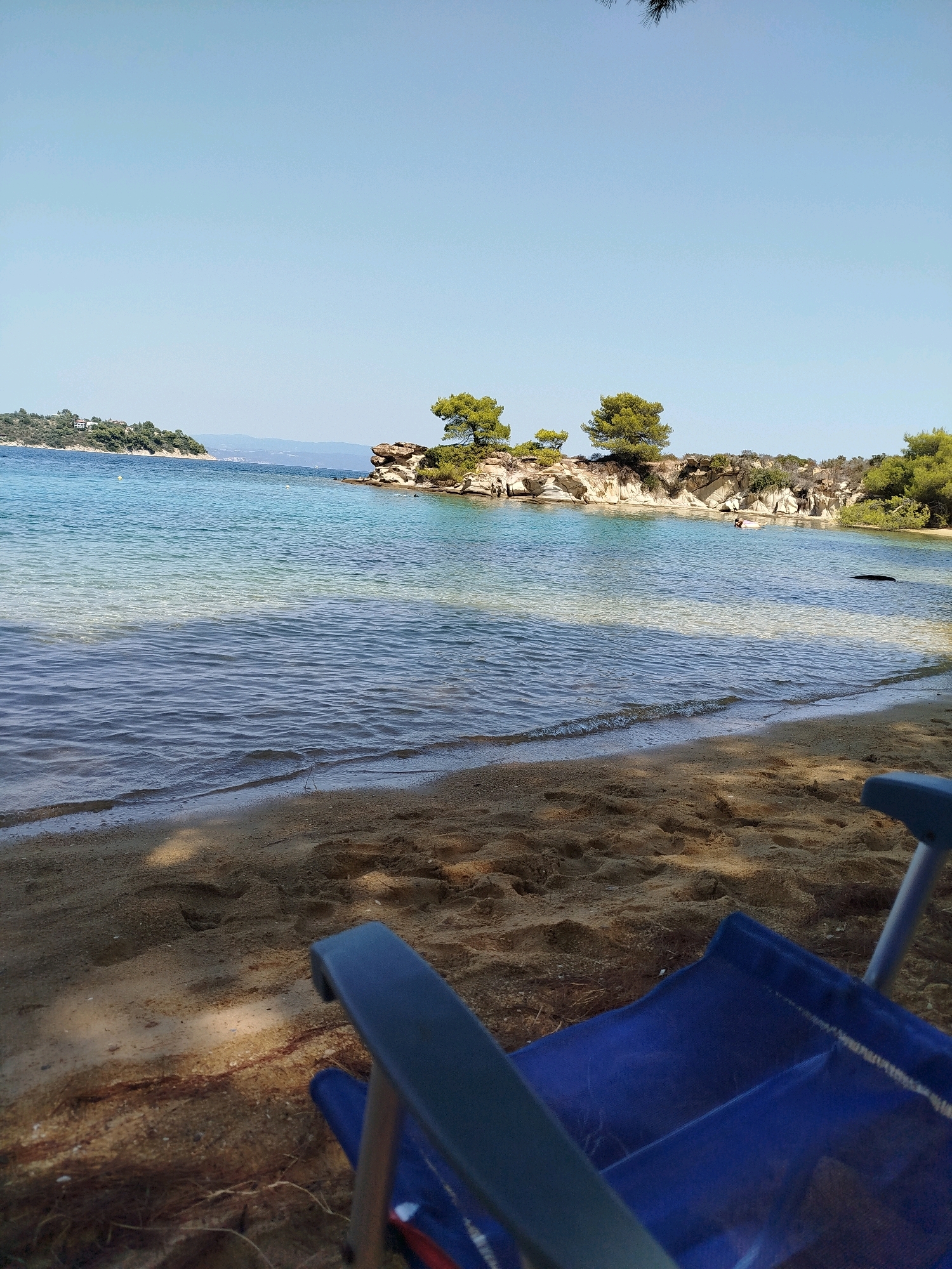 The image depicts a tranquil beach scene in Sithonia Municipal Unit, Greece. In the foreground, a blue beach chair is positioned on a sandy shore, with subtle shadows indicating sunlight filtering through foliage. The sand displays footprints, suggesting recent activity. The clear, turquoise water gently laps at the shore, revealing the sandy bottom.
In the mid-ground, a rocky outcrop adorned with evergreen trees juts into the sea. The water stretches towards a distant, verdant island with scattered buildings, bathed in bright sunlight under a clear, pale blue sky. The overall atmosphere is serene and inviting, characteristic of a Mediterranean coastal landscape.
