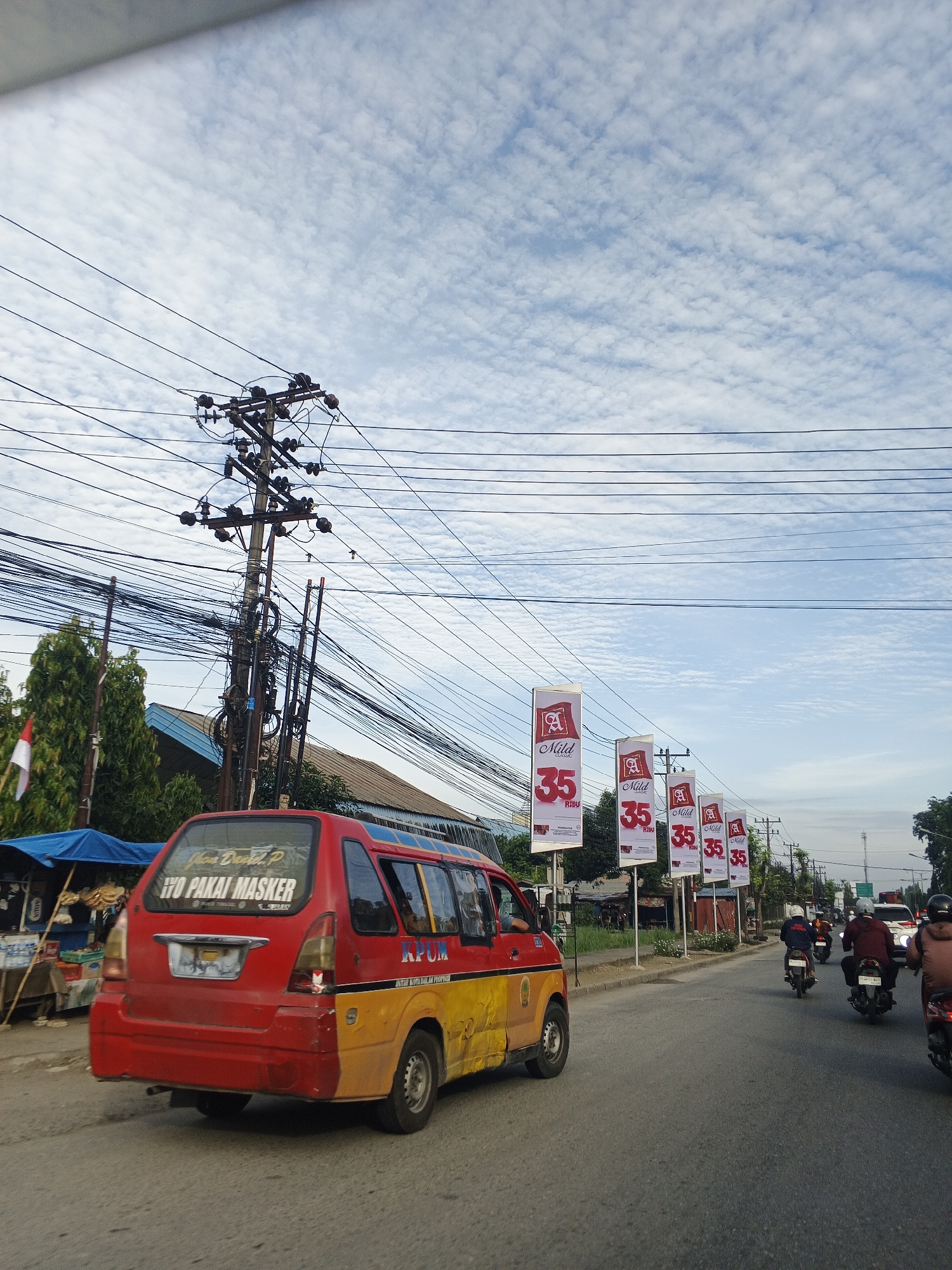 The image depicts a street scene in Tanjung Morawa, Indonesia, under a cloudy sky. In the foreground, a red and yellow van is traveling on the right side of the road. The van has text on its rear window that reads "IYO PAKAI MASKER", and "KPUM" is visible on its side. Several motorcycles with riders are also on the road, moving in the same direction as the van. Along the side of the road, a series of vertical banners display "A Mild 35 RIBU" graphics. A tall utility pole with numerous wires is prominent on the left side of the image, with trees and buildings visible behind it. A small market stall with blue tarpaulin is also present on the left. The overall scene suggests a typical urban or semi-urban street with vehicular traffic and advertising.