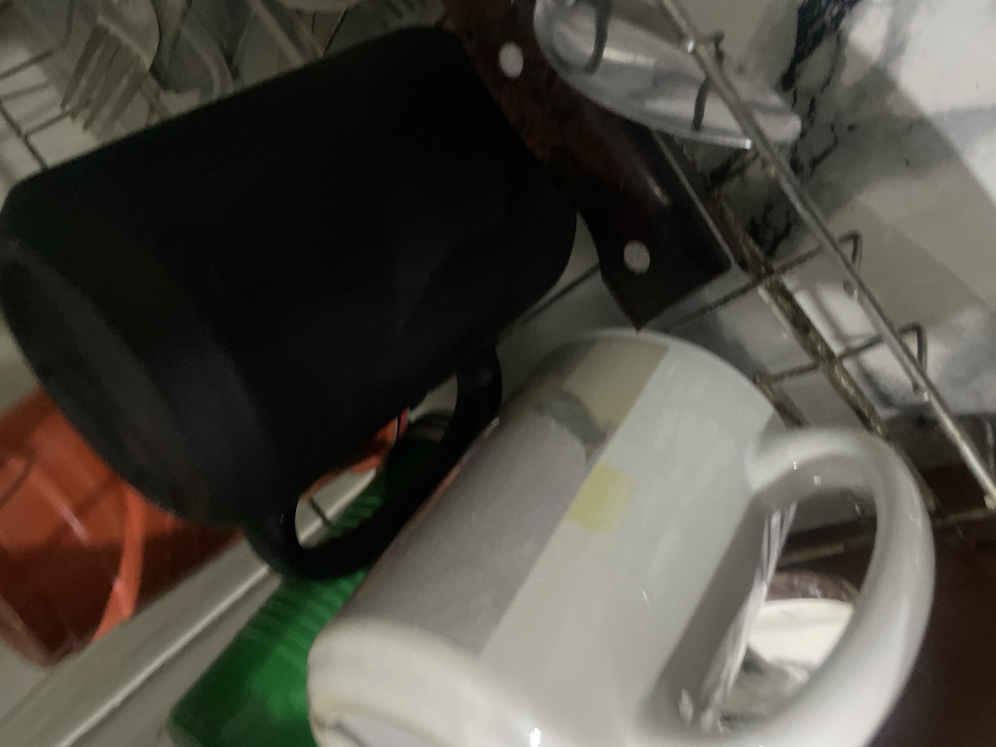 The image shows a close-up, tilted view of a kitchen dish rack with various items. A large black mug is in the foreground, angled towards the right. To its left and slightly lower is a white mug with a greyish-white exterior, its handle prominently visible. Behind these mugs, a red liquid is visible in a container, and a green object, possibly another container, is partially seen. A knife with a dark wooden handle is placed horizontally beneath the mugs, positioned on the dish rack. The overall environment appears to be a kitchen, with the dish rack being the central focus. The lighting is somewhat dim, with reflections visible on the surfaces of the mugs. There are no people or animals present in the image.