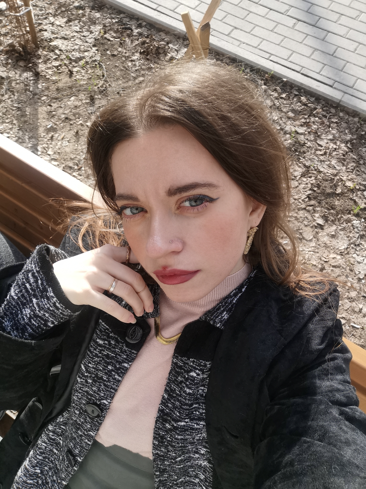 A woman is captured in a selfie-style shot, seated outdoors on a wooden bench under bright sunlight. She has long, dark brown hair, blue eyes accented with dark eyeliner, and wears red or maroon lipstick that appears slightly smudged on her upper lip. A small silver stud nose piercing is visible on her right nostril. Her expression is direct and intense, looking straight at the camera.
She is dressed in multiple layers: a light pink high-necked top, a black and white patterned, textured cardigan or sweater, and a black outer jacket. A gold-toned necklace with a snake-like chain is visible around her neck. On her left hand, which is held up near her chin, she wears a silver ring with small stones on her ring finger and a thin gold band on her pinky finger. Gold-toned, textured earrings are visible on both ears.
The background shows a ground covered with dry, light-colored leaves and bare soil, suggesting an outdoor, possibly park-like setting. A paved walkway or curb runs along the top right of the frame. The harsh sunlight creates distinct shadows, indicating a clear, sunny daytime. There is no visible text in the image.