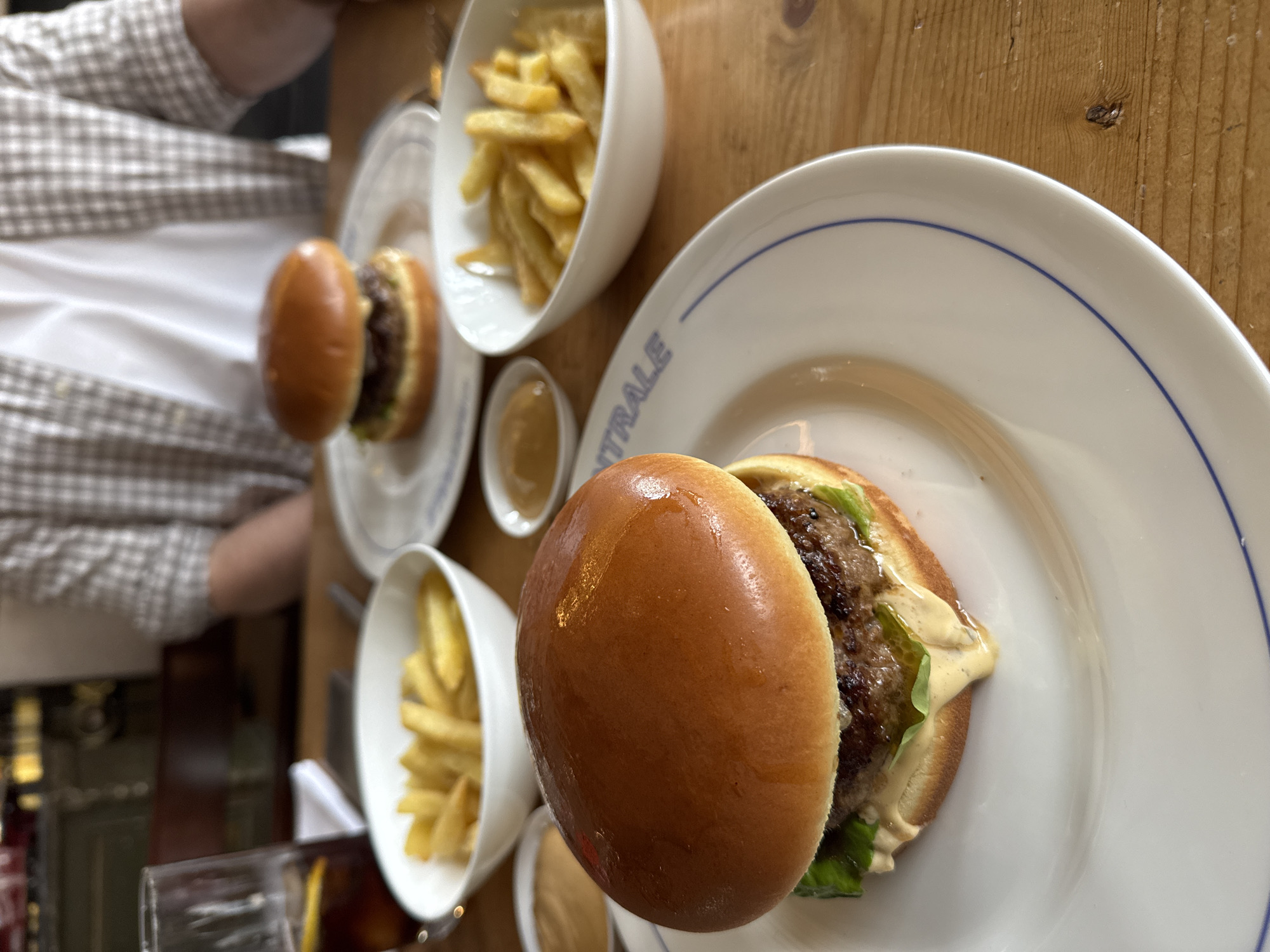 This image captures an indoor dining scene, likely in a restaurant or cafe in Radovići, Montenegro. The setting features a light-colored wooden table where two individuals are enjoying a meal.
In the foreground, a well-presented burger with a glossy bun, a beef patty, lettuce, and a creamy, light-colored sauce is positioned on a white plate. The plate has a blue rim and the word "CENTRALE" printed in blue text, likely indicating the establishment's name. Alongside this, a small white bowl is filled with golden French fries, and a tiny ramekin holds a light brown dipping sauce.
Further into the background, a second, slightly out-of-focus burger, plate, and bowl of fries are visible, indicating another diner. A person wearing a light-colored, plaid long-sleeved shirt is partially visible on the left side of the frame, largely obscured from the chest down. Another person's arm is subtly visible in the top left corner. A clear drinking glass is also present on the table.
The activity taking place is a casual meal, featuring classic burgers and fries. The lighting is consistent with an indoor setting, without strong natural light cues. The visible text "CENTRALE" provides a specific location cue for the dining establishment. The overall impression is a straightforward, pleasant dining experience.