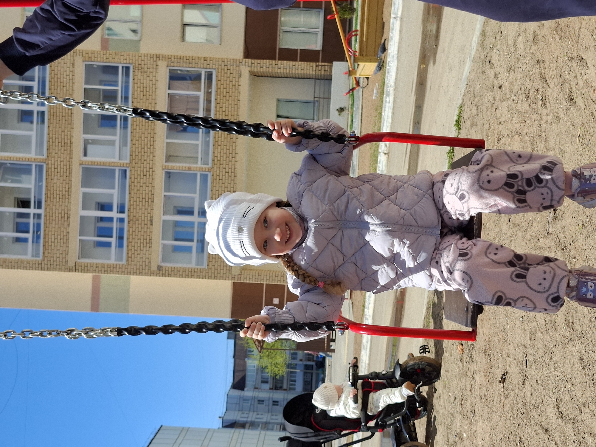 A child, wearing a white knitted hat, a light purple quilted jacket, and patterned light pink pants with teddy bear prints, is sitting on a red swing in a playground. The child is holding onto the black handles of the swing and smiling at the camera. Another person's arm is visible at the top left and top right corners of the image. Behind the swing is a beige and brown brick apartment building with several windows. In the foreground, to the right, there is sand and a tricycle with a baby wearing a white hat in it. The sky is blue. This scene is likely in the city of Chaikovsky, Russia.