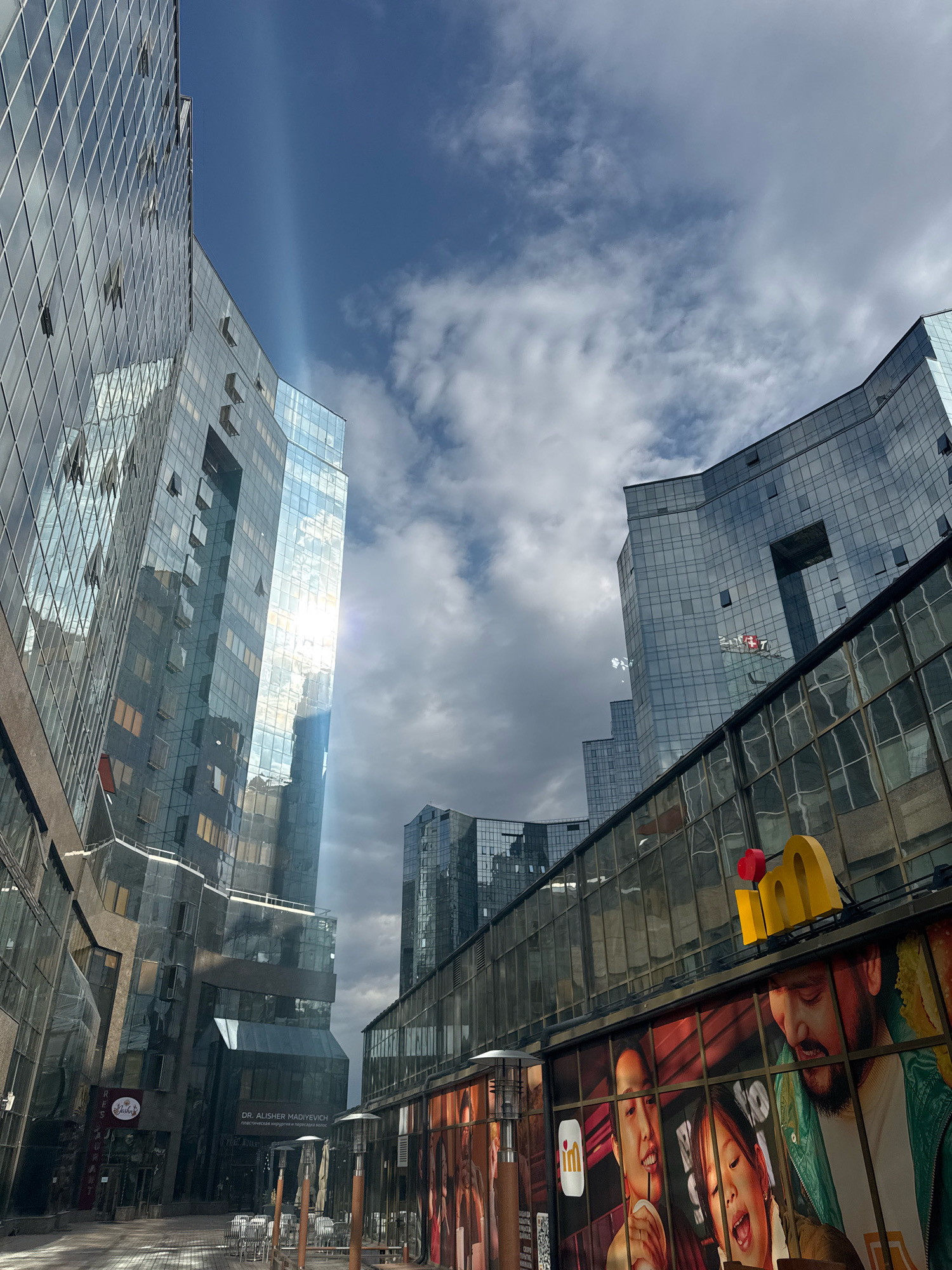 The image captures a street scene in Almaty, Kazakhstan, with modern glass-fronted buildings dominating the view. The sun shines brightly, creating a lens flare effect and casting beams of light through the clouds. In the foreground, a building with a large advertisement featuring people and a yellow "m" logo is visible. The advertisement appears to be for a fast-food restaurant. Across the street, the reflective surfaces of other glass buildings create a sense of depth and reflect the sky and surrounding architecture. The overall impression is one of urban development and contemporary design in Almaty.