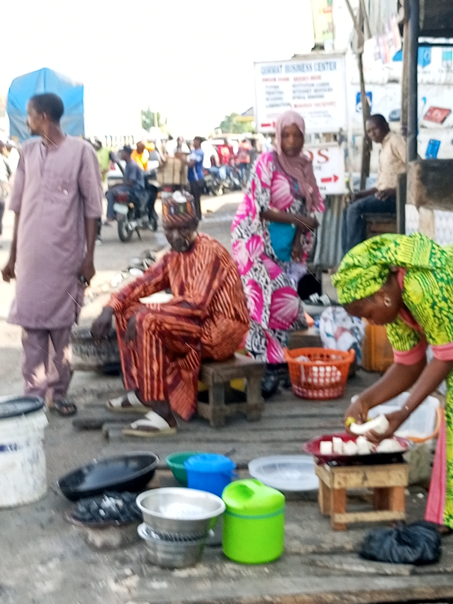 The image depicts a street scene in Gombe, Nigeria, likely a marketplace or commercial area. In the foreground, a woman in a vibrant green headscarf and dress is actively preparing food. She is holding a peeled banana and placing pieces onto a red platter situated on a small wooden stool. Next to her, an orange plastic basket sits on the ground, containing white food items. To the left, a man in a traditional orange and red striped attire with a cap is seated on a stool, looking towards the food preparation. A man in a light purple traditional outfit stands behind him, looking away from the camera. In the background, other individuals and a motorcycle are visible, suggesting a busy urban environment. A white sign with text is mounted on a pole in the mid-ground, and a blue tarp is visible further in the distance. The ground appears to be paved or made of wooden planks. The overall atmosphere suggests an outdoor food vending or preparation area.