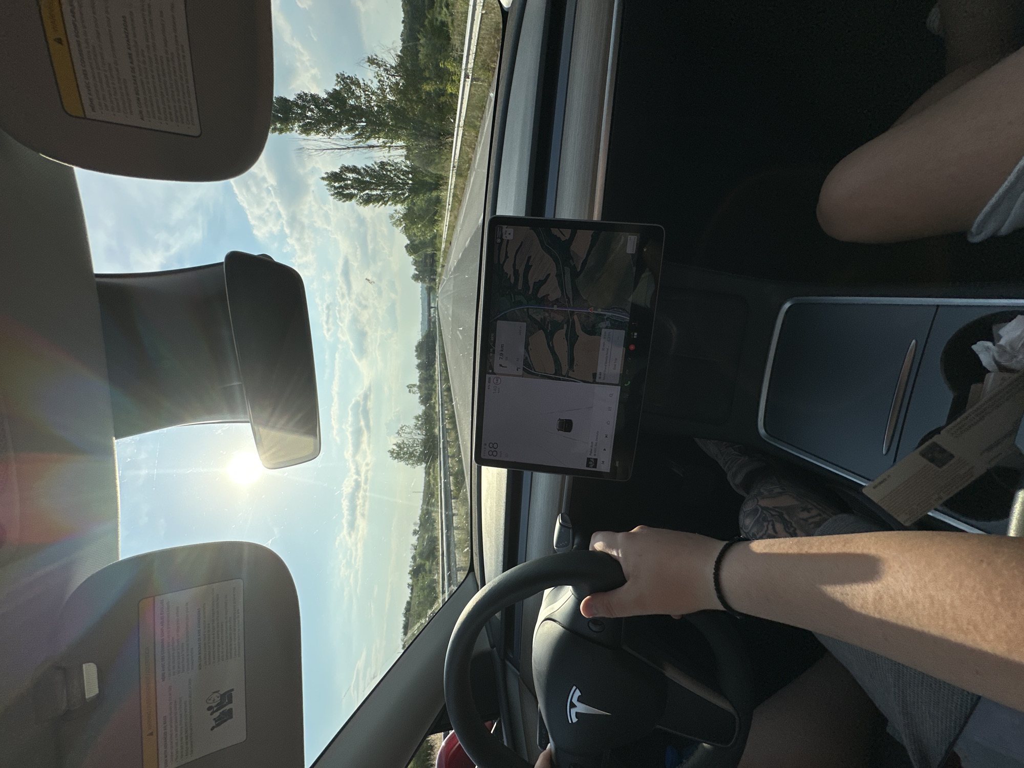 The image provides a driver's perspective from within a moving Tesla vehicle on a highway near Kapitan Andreevo, Bulgaria, under bright daytime conditions. The driver's hands grip the steering wheel, with the right arm, adorned with a black braided bracelet, prominently visible. Tattoos are discernible on the driver's left leg, partially covered by light-colored shorts. A passenger's right knee is visible in the lower right foreground. Both driver and passenger sun visors are in the upright position, displaying multi-language warning labels. The central Tesla touchscreen displays a speed of "88", a navigation map indicating a route with 7.9 km remaining, an outside temperature of "14 C", and an Autopilot visualization showing the car within its lane with a detected vehicle ahead. A media player interface is partially visible at the bottom of the screen. The center console contains an open storage compartment with crumpled paper or tissues. Through the windshield, a multi-lane asphalt highway stretches into the distance, flanked by green hills, trees, and guardrails. Tall, slender trees are visible along the right side of the road. A bridge or overpass is discernable on the distant horizon. The sky is blue with scattered white clouds, and the low sun is directly ahead, casting bright glare and lens flares across the windshield.