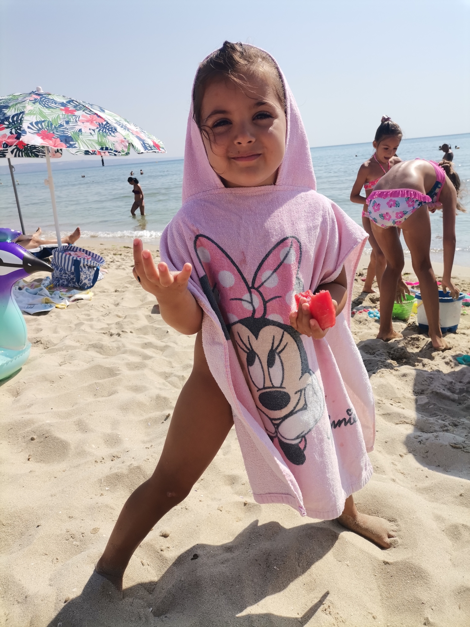 In Kranevo, Bulgaria, on a sandy beach, a young girl wearing a pink hooded poncho with a Minnie Mouse design stands in the foreground. She is holding a piece of watermelon in her right hand and looking at the camera with a smile. Behind her, other children are visible near the water's edge, with a colorful beach umbrella and various beach items scattered on the sand. The sea stretches out to the horizon under a clear blue sky.