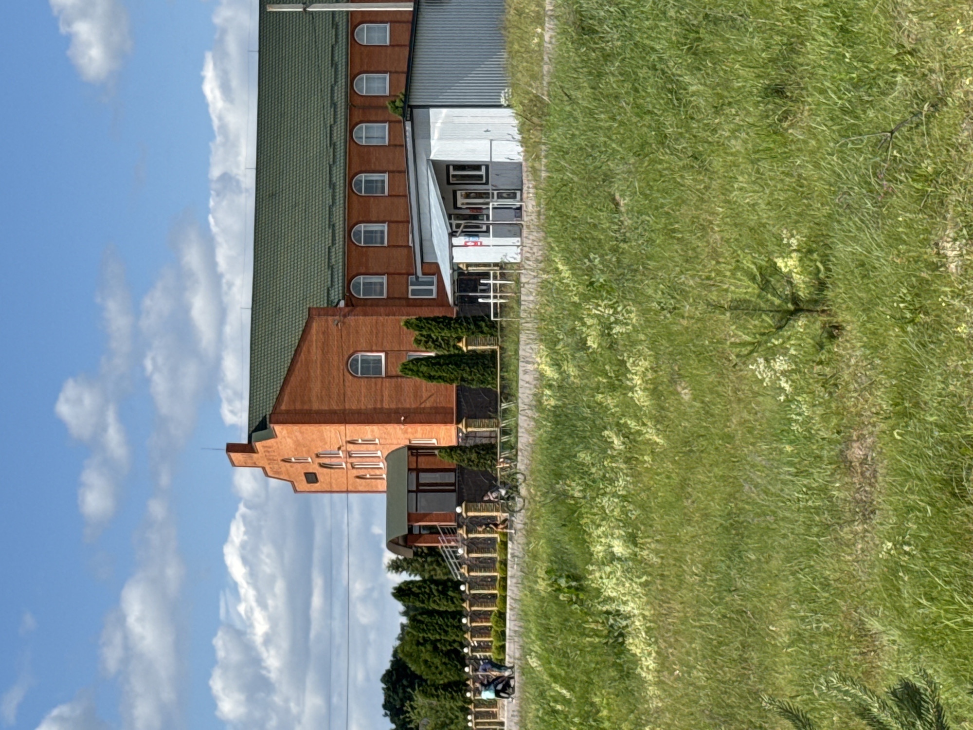 The image depicts a side view of a multi-story building with a red brick facade and a green roof. The building has several arched windows on its upper levels. Adjacent to the building is a paved walkway, bordered by a fence and a lush, green grassy area. A bicycle is leaning against the fence. In the background, more trees and vegetation are visible. The sky is blue with scattered white clouds, suggesting it is daytime with fair weather. The overall scene appears to be a quiet outdoor setting, possibly a residential area or a commercial establishment with outdoor space.