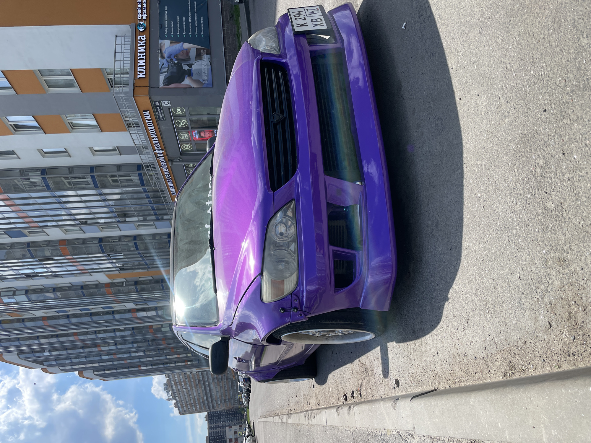 The image displays a bright purple, heavily modified car, specifically a Lexus IS 200/300 (or Toyota Altezza), parked on a paved street in the city of Unknown, Russia. The car, occupying the foreground, features wide-body fender flares, an aggressive front bumper with large air intakes, and an aftermarket grille with a black emblem. Its license plate, "K 294 XB 147", is affixed to the right side of the front bumper. Silver multi-spoke wheels are visible, with the front wheel angled towards the viewer. A strong sun glare obscures part of the windshield and hood.
In the mid-ground and background, modern, tall residential or commercial buildings dominate the urban landscape under a partly cloudy blue sky. The building directly behind the car features alternating sections of reflective glass facades and white/orange walls. On the ground floor of this building, a business sign in Cyrillic letters reads "КЛИНИКА СЕМЕЙНОЙ ОФТАЛЬМОЛОГИИ" (Family Ophthalmology Clinic), with the address "Екатерининская 3/4" also visible. An advertisement for the clinic, depicting an eye examination, is displayed on the building's exterior. Another building, slightly further back and to the left, is also visible. The street surface is asphalt, with a concrete curb lining the left side. Shadows from the car and buildings are cast onto the street, indicating a sunny day. No individuals are visible in the scene.