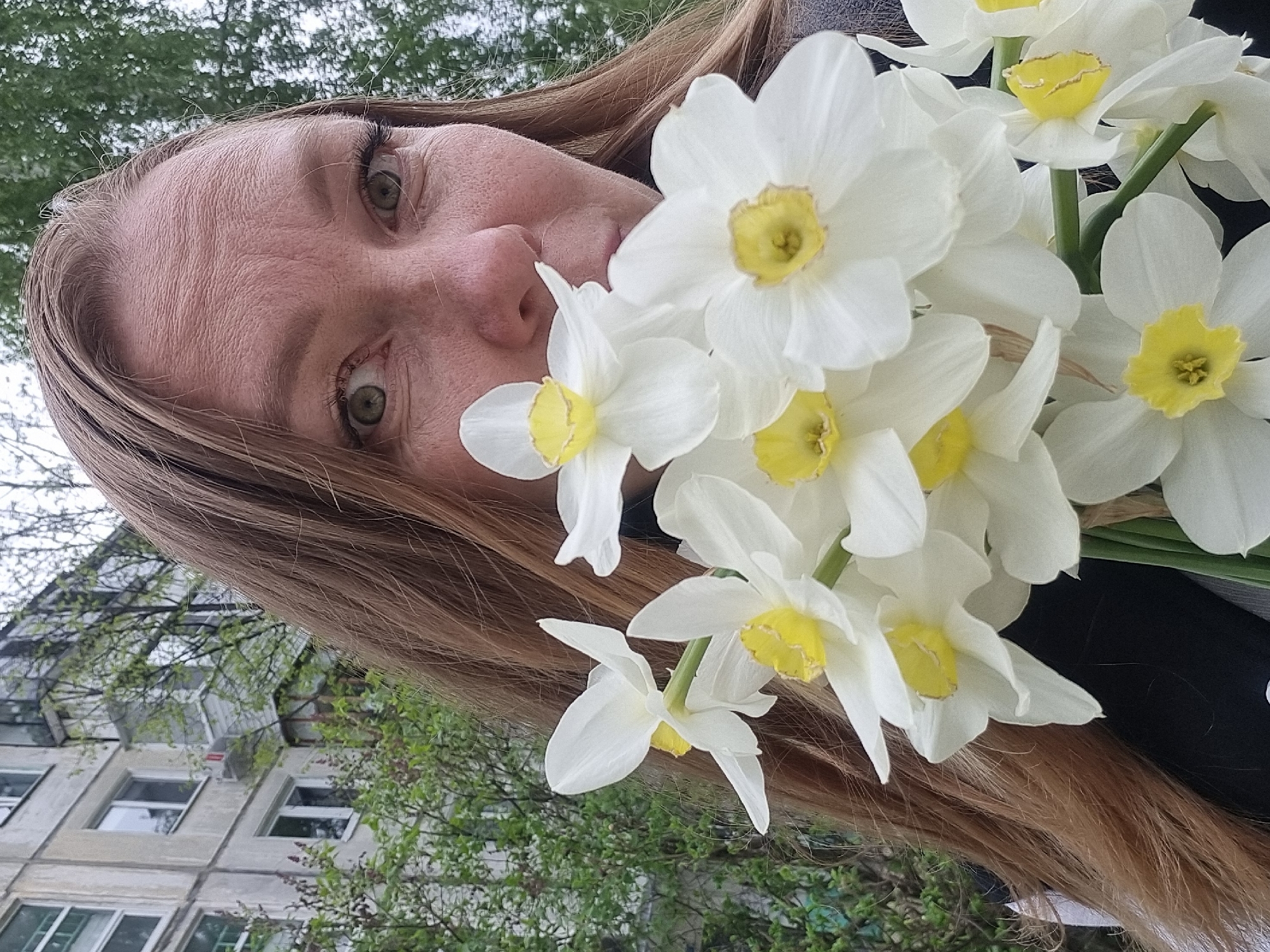 A woman with light brown hair and hazel eyes is holding a bouquet of white and yellow daffodils in front of her face. She is outside with a building visible in the background. The location is Chaikovsky, Russia. The sky is overcast.