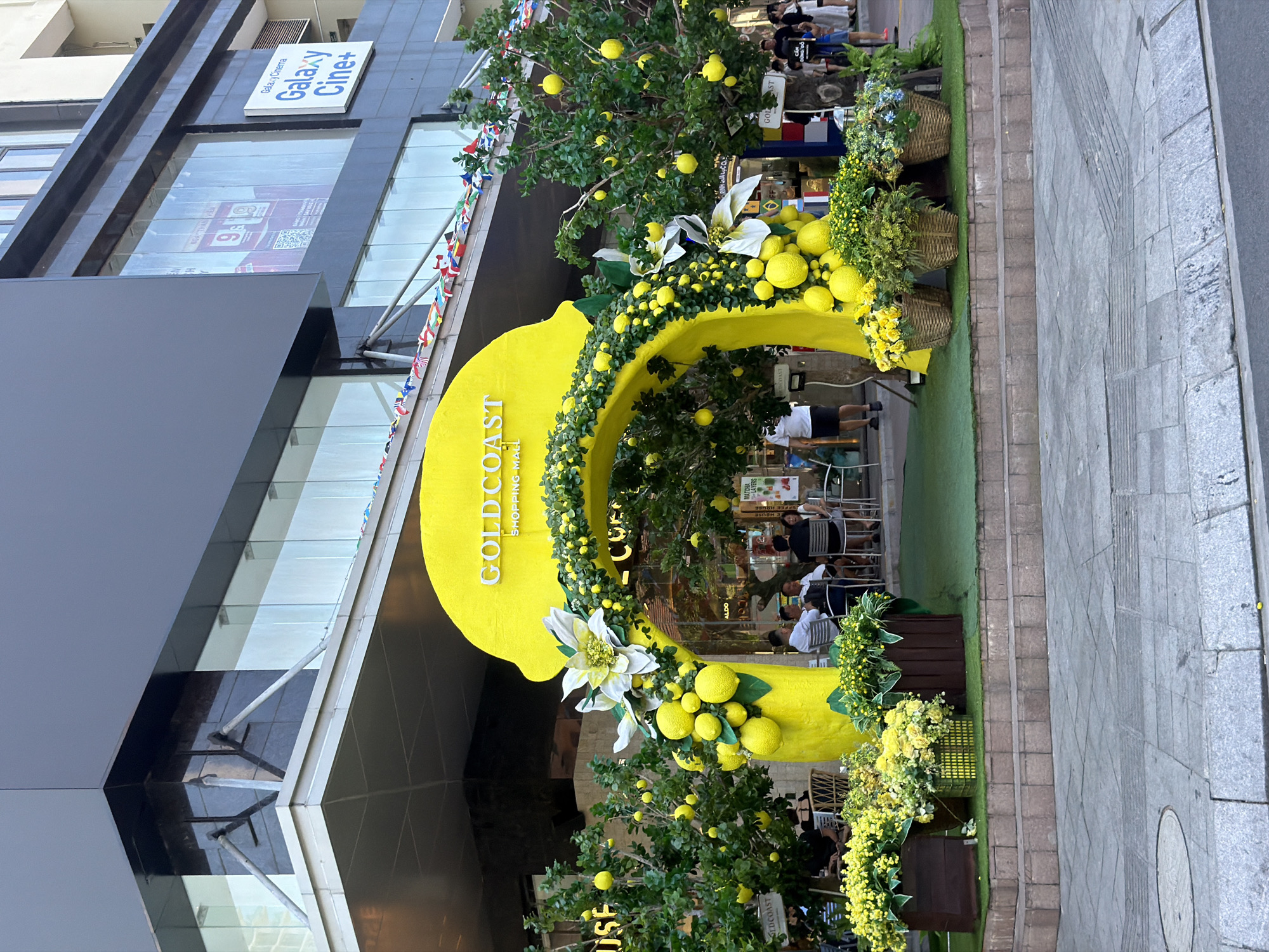 In the city of Nha Trang, Ward, Vietnam, a vibrant yellow archway decorated with artificial lemons and white flowers stands prominently outside the Gold Coast Shopping Mall. The archway is flanked by trees also adorned with artificial lemons. In front of the arch, several potted plants filled with yellow and green foliage are arranged on tiered platforms covered with green artificial turf. Behind the arch, a group of people are seated at tables, appearing to be dining or relaxing. The background shows the modern facade of the Gold Coast Shopping Mall, with its glass windows and signage for "Galaxy Cinema". A street with pedestrian walkways is visible in the foreground.