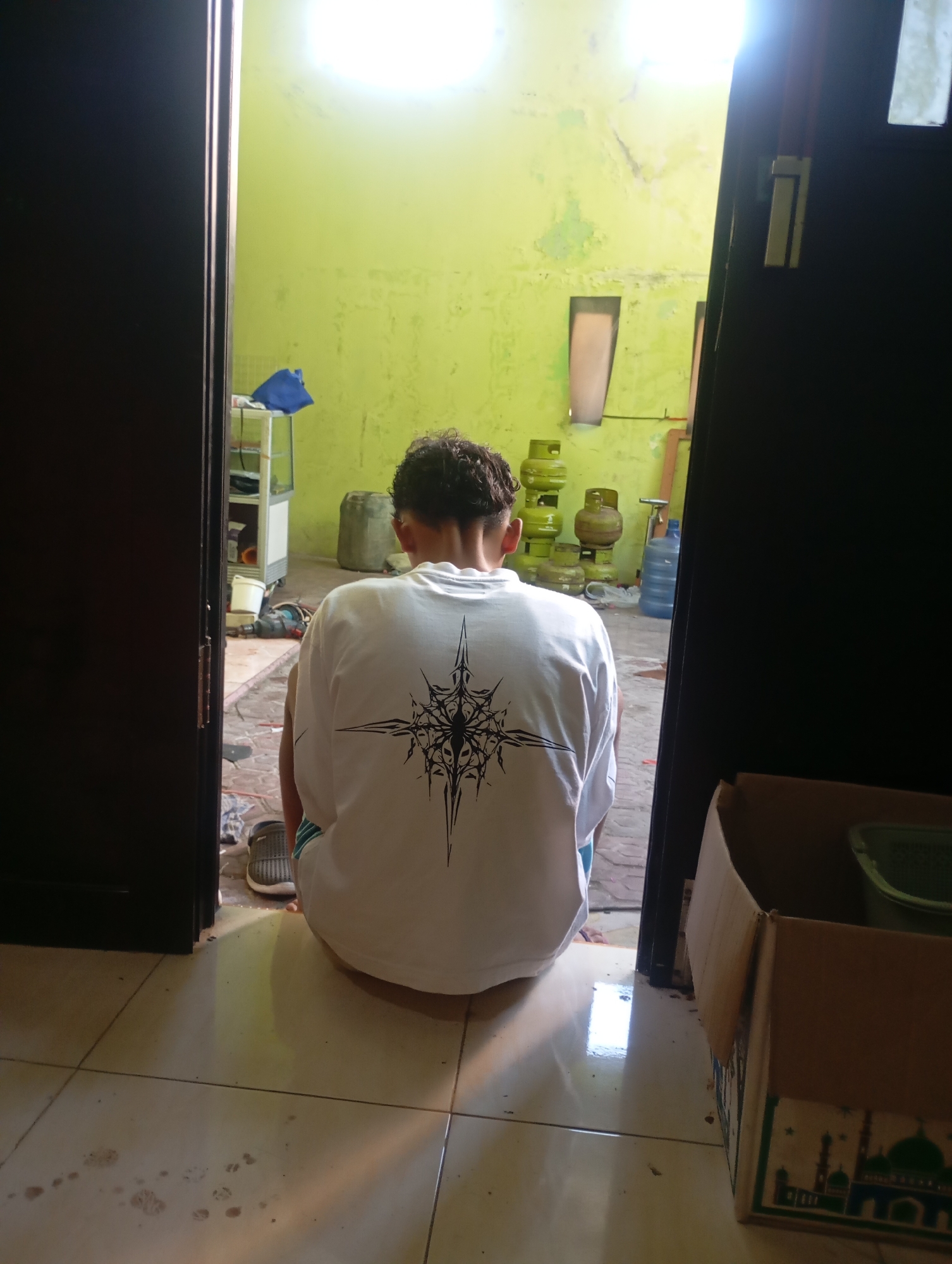 The image depicts the rear view of a person sitting on a tiled floor, facing away from the camera. The person is wearing a white t-shirt with a black graphic design on the back. They are also wearing blue shorts and dark-colored shoes. The setting appears to be an interior space with a vibrant lime green wall. In the background, several green propane tanks are visible, along with a large blue water jug. To the left, there is a shelf unit with some items on it, and a dark blue cloth. To the right, a cardboard box with Islamic architectural designs is prominent. The floor is light-colored tile with some apparent water stains and paw prints. The overall scene suggests a domestic or storage area in Tukmudal, Indonesia, likely a residential setting given the presence of household items and the open doorways suggesting a transition to an outdoor or semi-outdoor space. The lighting suggests natural light coming from the left and a bright overhead light source.