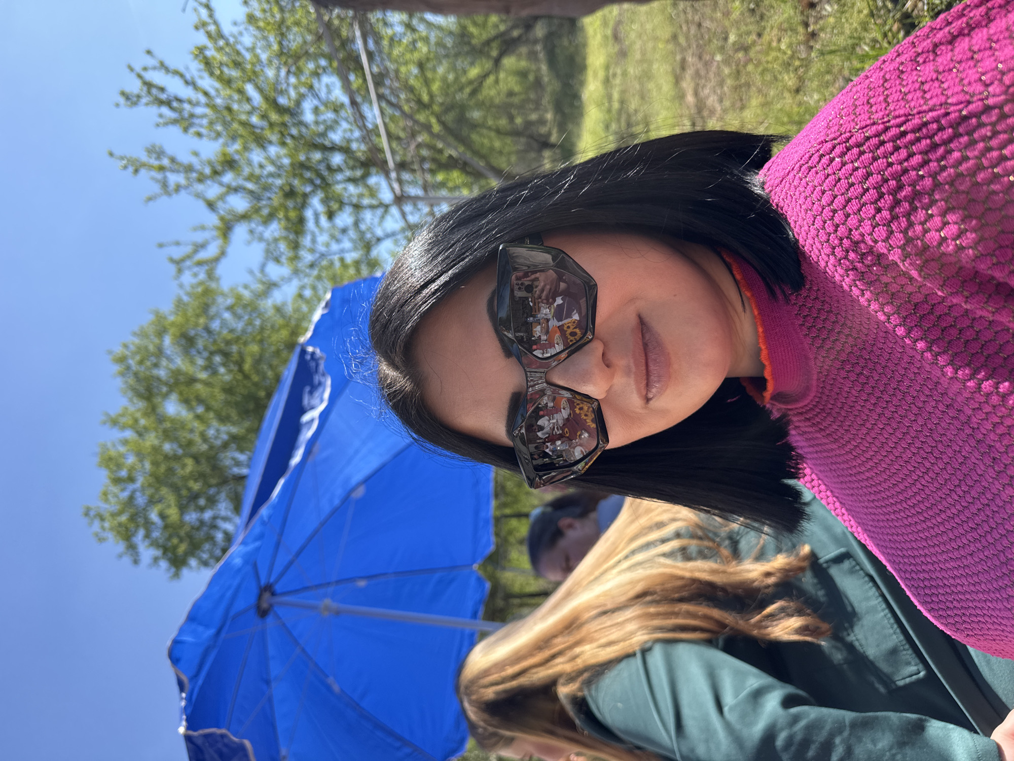 A woman with dark hair and sunglasses poses in front of a bright blue umbrella, likely in an outdoor setting near Lom, Bulgaria. The reflection in her sunglasses shows a crowded table with food, flowers, and people. She wears a textured pink sweater and stands next to a person with long blonde hair wearing a dark green jacket. The background features a tree and a clear blue sky.