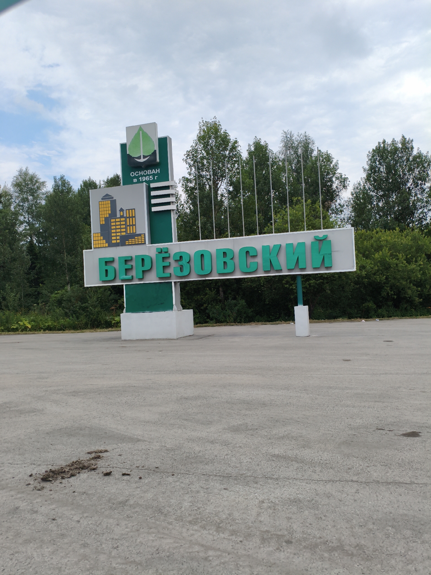 A large, white sign with green lettering reads "БЕРЁЗОВСКИЙ" (Beryozovsky) in the foreground. Above this, a smaller sign shows a stylized green leaf and an anchor, with the text "ОСНОВАН В 1965 Г" (Founded in 1965). To the left of the main sign is a depiction of yellow buildings. The sign is supported by a green and white structure. The sign stands on a paved area, possibly a road or parking lot, with a patch of dirt and debris to the left. Behind the sign, a line of trees forms a backdrop under a cloudy sky. The overall scene is outdoors and appears to be an entrance to the city of Beryozovsky, Russia.