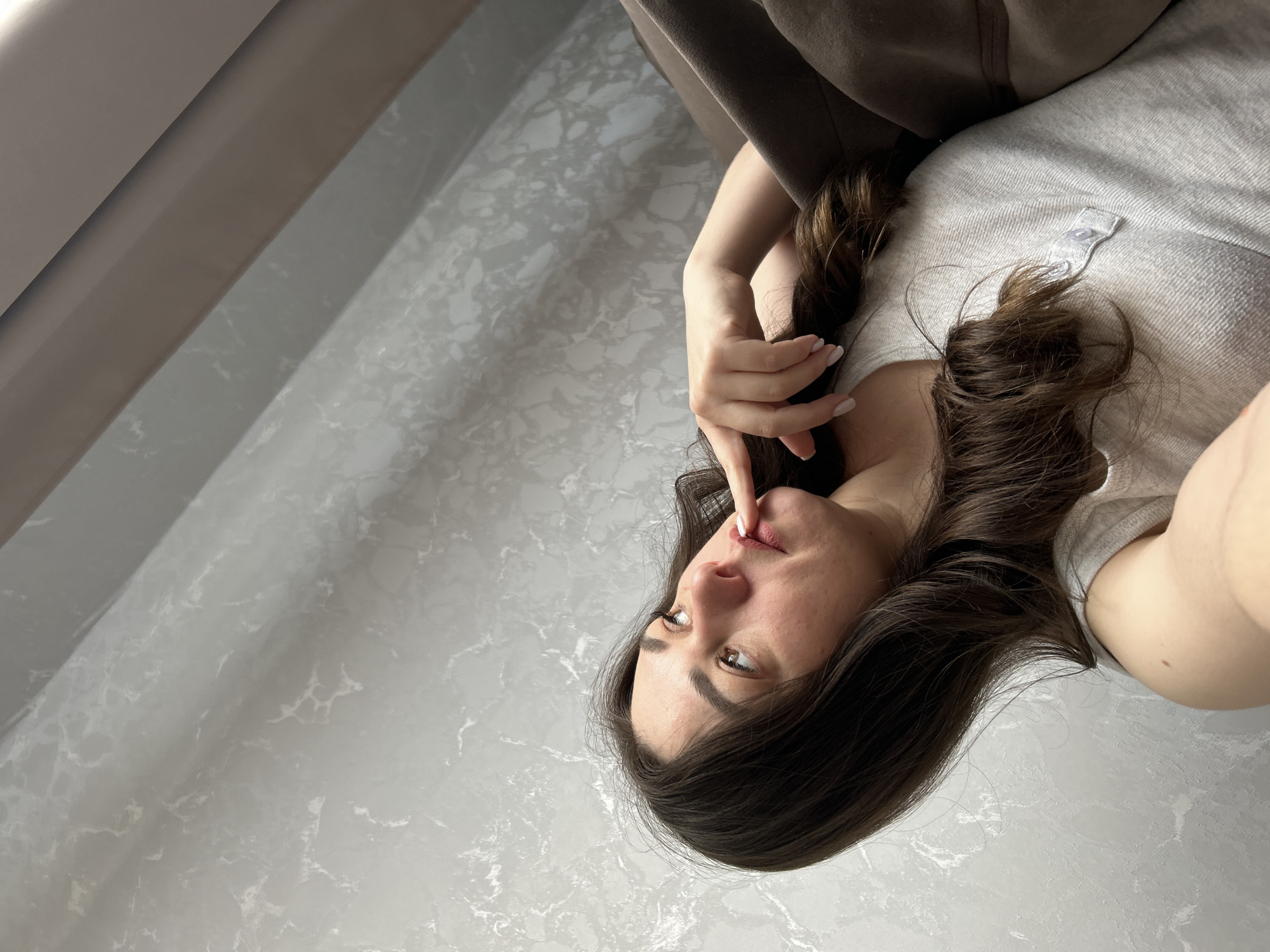 A woman with long, dark brown hair is lying down. She is wearing a light gray shirt and a brown garment drapes over her. Her right hand is raised, and her index finger touches her upper lip. The background consists of a gray patterned wall and a light-colored surface. There is no information to determine a specific location in Chaikovsky, Russia.