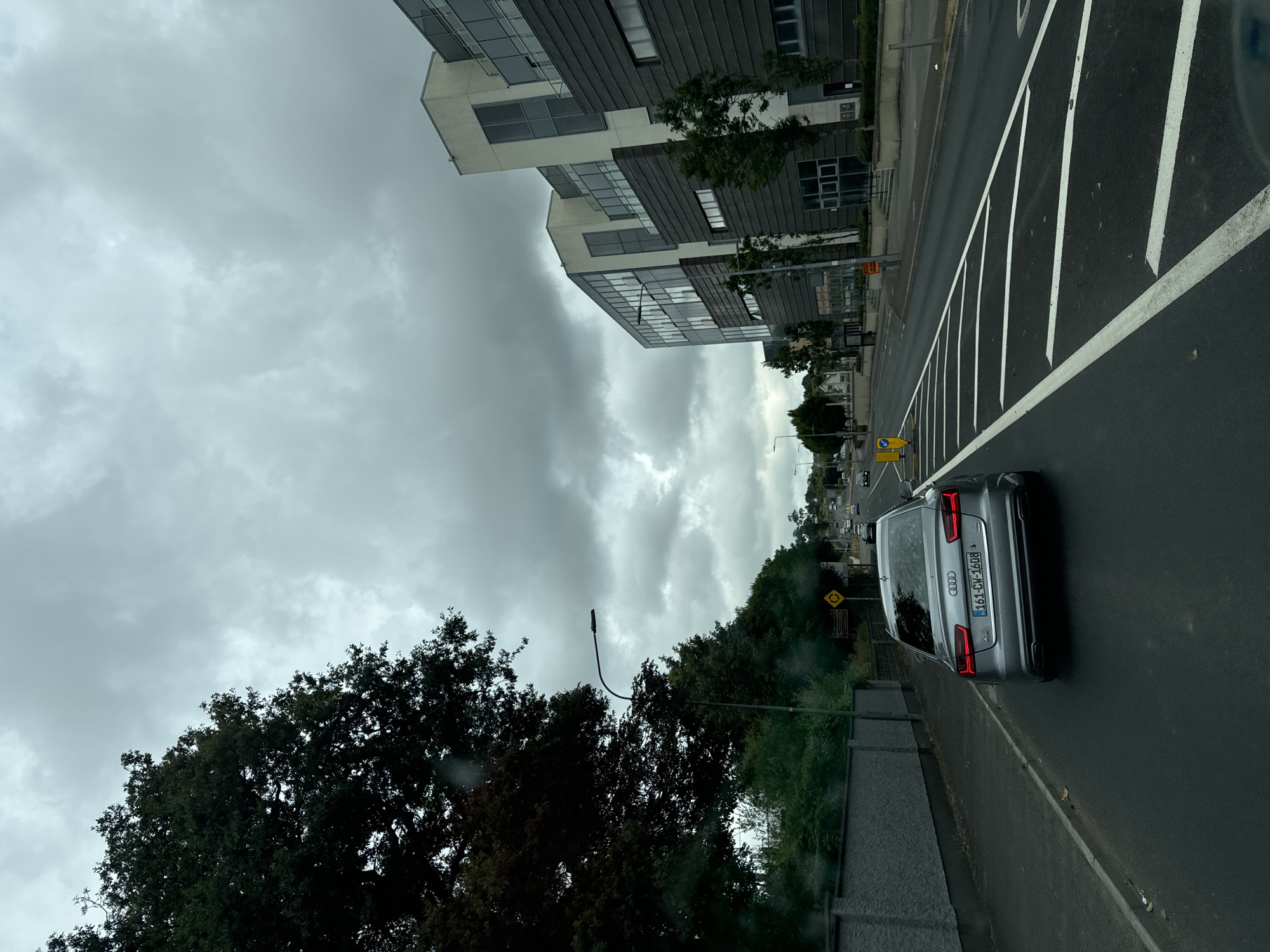 The image shows a grey Audi A6 car with the license plate "161-CW-1608" driving on a road in Waterford, Ireland. The car is in the foreground, and the road appears to be wet, suggesting recent rain. On the right side of the road are modern office buildings. On the left side of the road, there is a grey wall, a sidewalk, and trees. The sky is overcast with grey clouds. The overall scene depicts a typical urban environment in Ireland.
