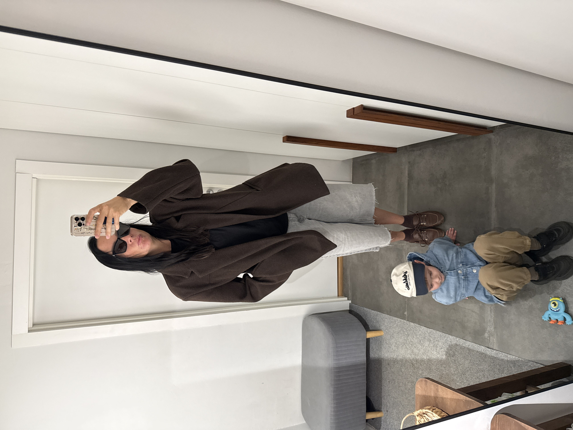 In a mirrored reflection of a modern interior, a woman stands taking a selfie with her phone. She is wearing a dark brown coat over a black top and light gray pants. She has on black sunglasses and her dark hair is long and styled. In the foreground, a young child is lying on the floor. The child is wearing a light blue denim jacket over a tan shirt and tan pants, along with a white and blue baseball cap. To the right of the child, a small blue toy monster is visible. The setting appears to be indoors, with a gray concrete floor and white walls. There is a gray bench with wooden legs to the left of the child and a wooden shelf structure in the lower right corner. The lighting suggests it could be daytime. The image is oriented vertically. The text on the child's cap is "CHAMPION".
