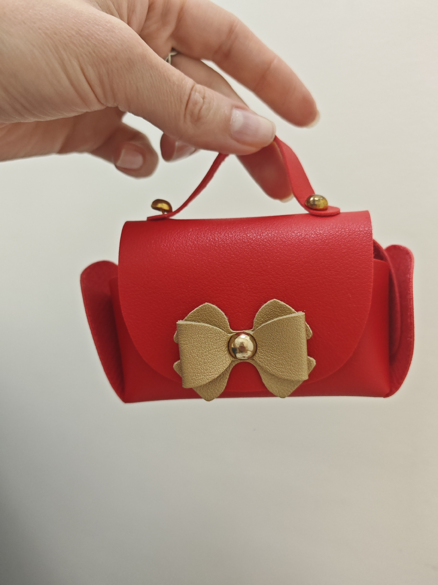 A hand holds a small, red, purse-shaped object with a gold bow and gold hardware against a plain white background. The purse has a folded flap closure and a small handle. The hand is positioned above and behind the purse, with the fingers gently holding the handle. The background is evenly lit and featureless. The location is Unknown, Unknown.