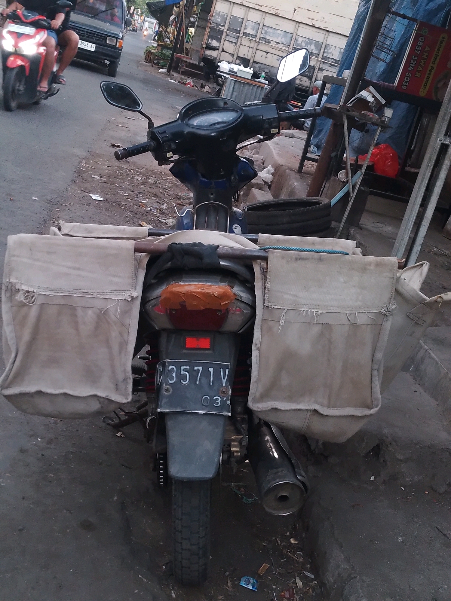 A motorcycle with large saddlebags is parked on a paved street in Kertosono, Indonesia. The motorcycle is dark blue with a black seat and a silver exhaust pipe. The saddlebags are made of beige canvas and appear to be old and worn. The motorcycle has a license plate that reads "B 3571 V". In the background, there is a black car, a red scooter, and a building with a blue tarp. The street is lined with debris and there is a red plastic bag on the ground. The overall scene depicts a typical street in a developing country, with a mix of vehicles and a somewhat unkempt environment.