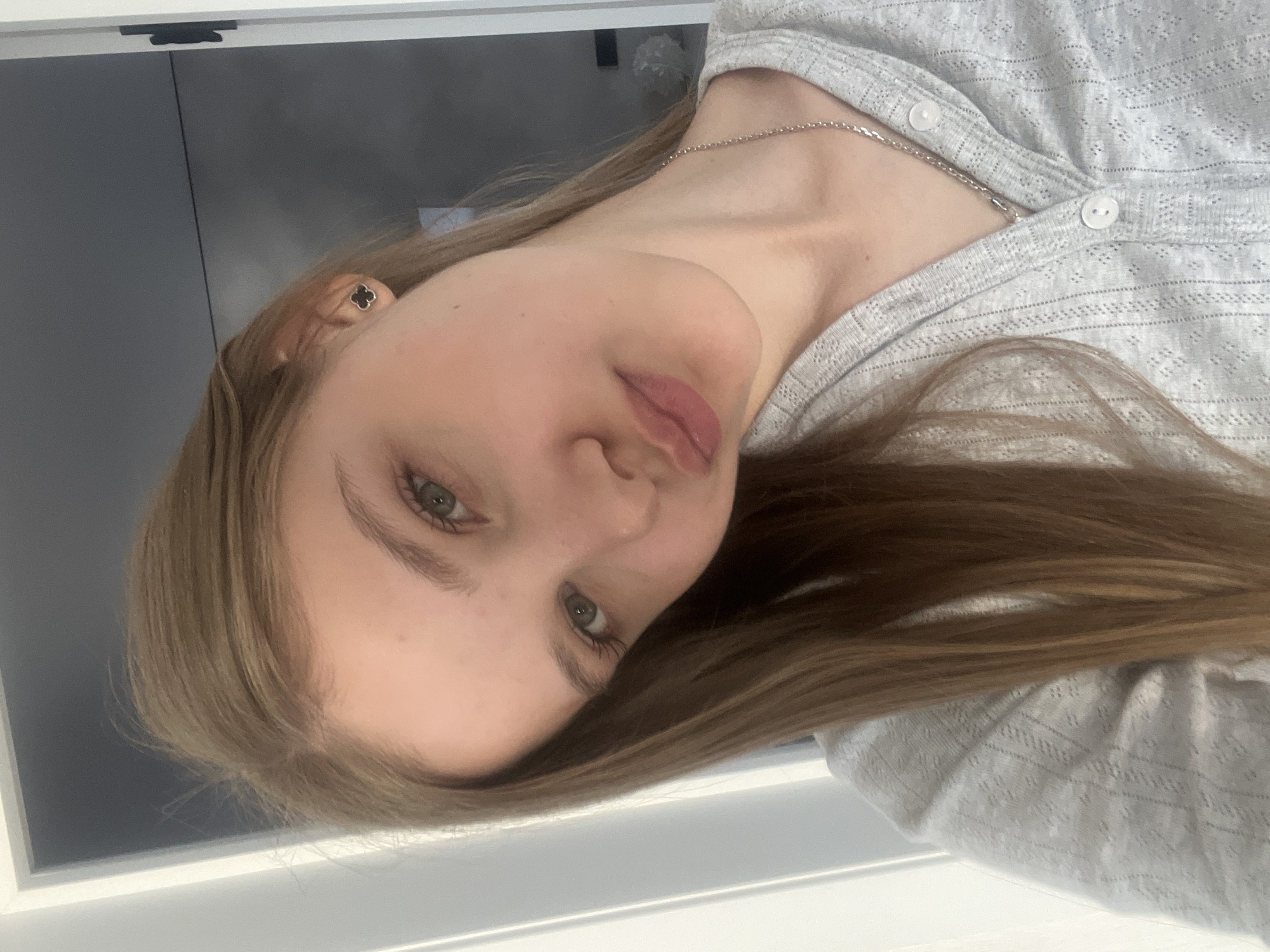 A portrait-style photograph features a young woman with long, light brown hair and light green or hazel eyes, looking directly at the camera. She has fair skin and her lips are lightly tinted pink. She wears a light grey top with a textured, possibly open-knit, pattern, and a slender silver chain necklace. A small, black, four-leaf clover-shaped stud earring is visible in her left ear. The background consists of a plain dark grey surface, likely a wall, with bright white horizontal borders at the top and bottom edges of the frame, suggesting an indoor setting. The lighting is bright and even, indicating daytime. Her expression is neutral and calm. While the location context provided is Votkinsk, Russia, there are no specific visual cues in the image to confirm this. No text is visible.