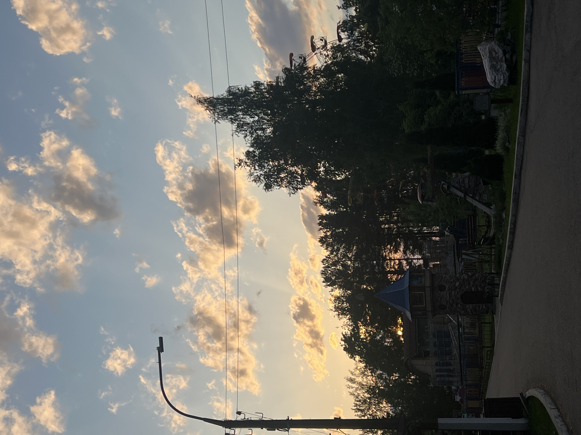 The image depicts an outdoor scene, likely a park or recreational area in the city of Chaikovsky, Russia. It shows a playground partially obscured by trees, with a castle-like structure and other play equipment visible. The sky is filled with clouds, illuminated by the setting sun, creating a warm, golden light. Power lines run across the sky, and a curved lamppost is visible in the foreground. The scene is captured at an angle, suggesting a street or walkway runs along the right side of the frame.
