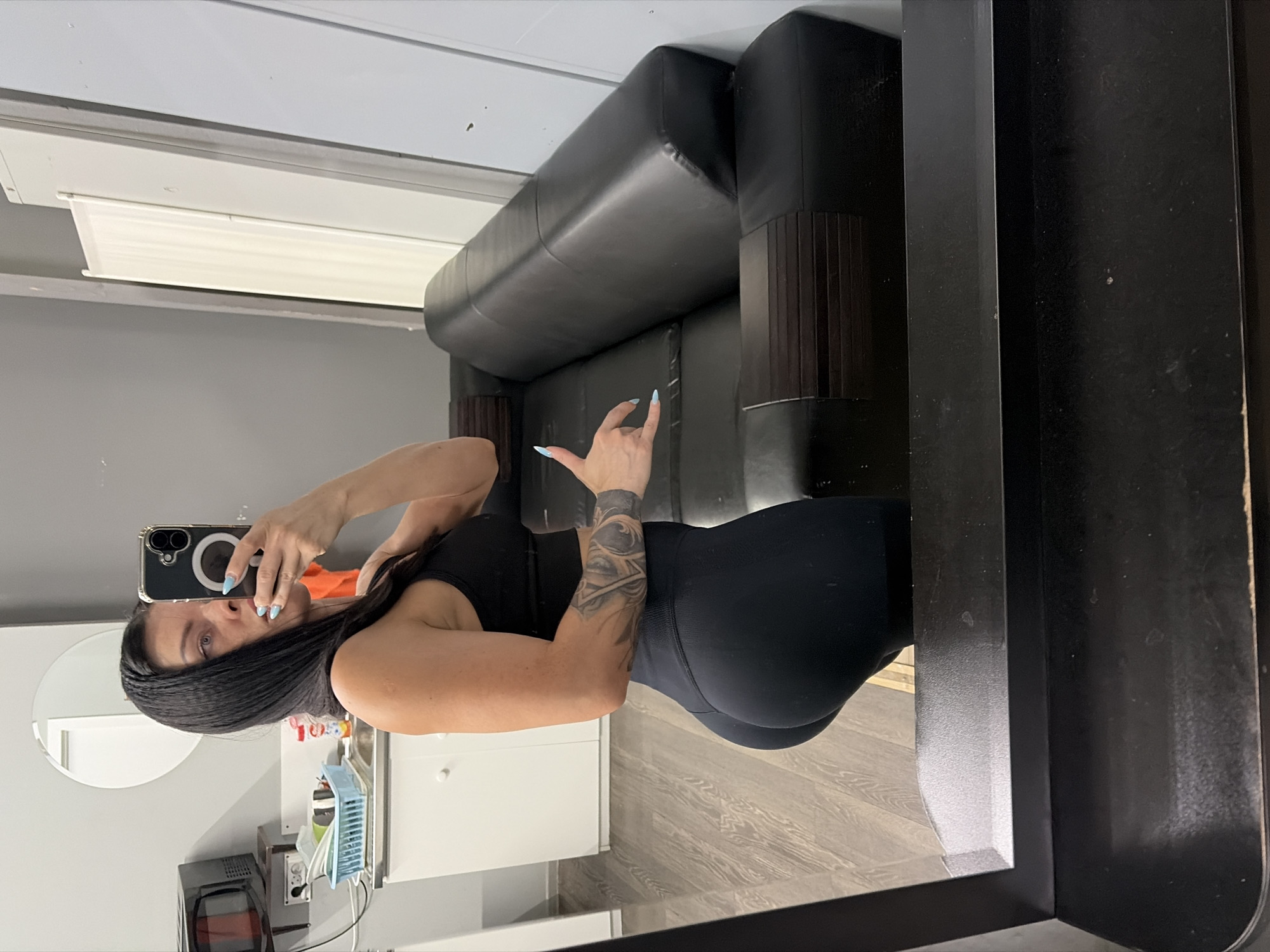 A woman is taking a mirror selfie in a room with grey walls and light wood flooring. She is wearing a black sports bra and black leggings, and has a tattoo on her left arm. In the background, there is a black leather couch and a set of white cabinets. There is also a microwave on a shelf and a sink with a blue basket of items. The scene appears to be in a residential setting, possibly an apartment or house. There is no information to indicate the location is in Ufa, Russia.