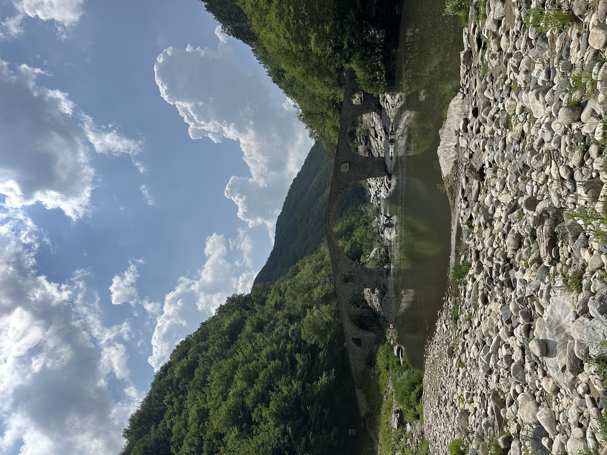 The image presents a serene natural landscape in Diadovtsi, Bulgaria, featuring an ancient stone arch bridge crossing a calm river. The view is from a rocky riverbank, looking across the water towards the bridge and the verdant, forested mountains beyond.
The historic bridge, constructed from robust grey stones, exhibits multiple arches gracefully spanning the tranquil river. At least three distinct arches are visible, their elegant curves creating striking reflections in the dark, reflective green water below. The bridge appears well-integrated into the natural environment, suggesting a long history.
Beyond the bridge, steep mountains rise, densely covered in a vibrant canopy of green trees and lush foliage, extending high into the distance. The sky overhead is a clear, bright blue, dotted with scattered white and light grey cumulus clouds that add depth to the expansive heavens.
In the immediate foreground, to the right, a wide bank of the river is covered with numerous smooth, rounded stones and pebbles, varying in size and tone. Sparse patches of bright green grass sprout intermittently among the rocks. The river's surface is remarkably still, accurately mirroring the sky, the surrounding trees, and the arches of the bridge, enhancing the scene's peaceful ambiance.
The image captures a beautiful, undisturbed natural setting, likely on a sunny day during late morning or early afternoon, indicated by the clear lighting and distinct cloud formations. There are no people or discernible text visible in the frame.