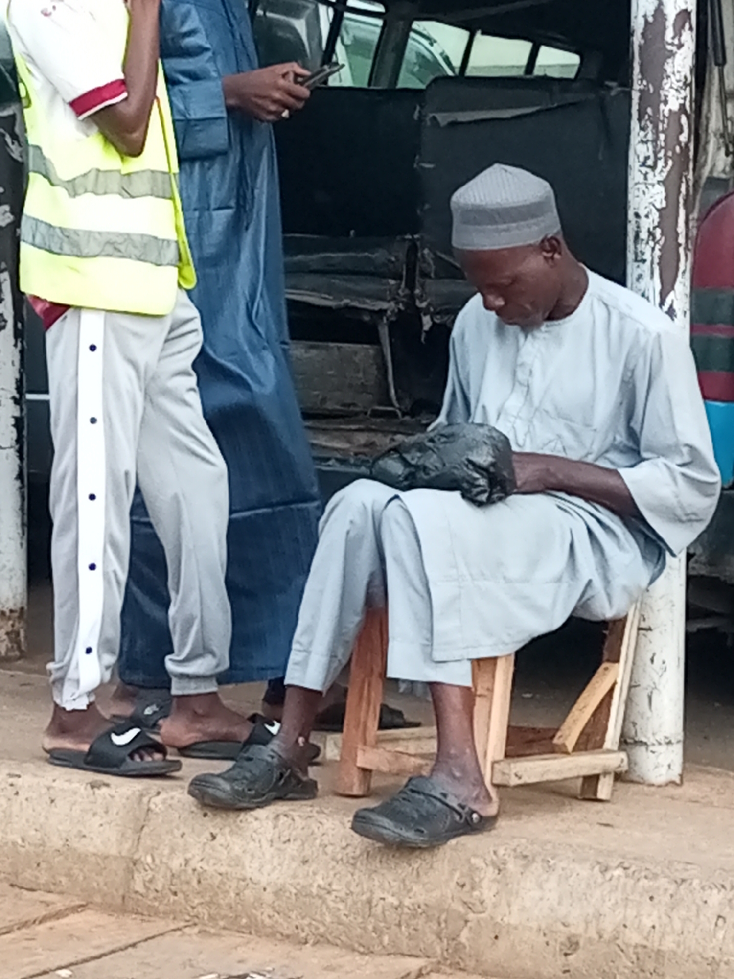 The image shows three people standing and sitting on a textured, uneven concrete ledge in what appears to be an outdoor urban setting in Gombe, Nigeria. The person on the left is wearing a white shirt with red trim, a yellow high-visibility vest, and light gray track pants with white stripes and blue buttons. They are holding a mobile phone. To their right, a person in a long, flowing blue garment stands, also appearing to be holding a phone. In the foreground, a man seated on a wooden stool wears a light blue traditional outfit and a grey skullcap. He is holding a dark, lumpy object wrapped in plastic in his lap. The background is out of focus, showing what looks like a vehicle and a distressed painted wall. The overall scene is casual, possibly a street corner or market area.