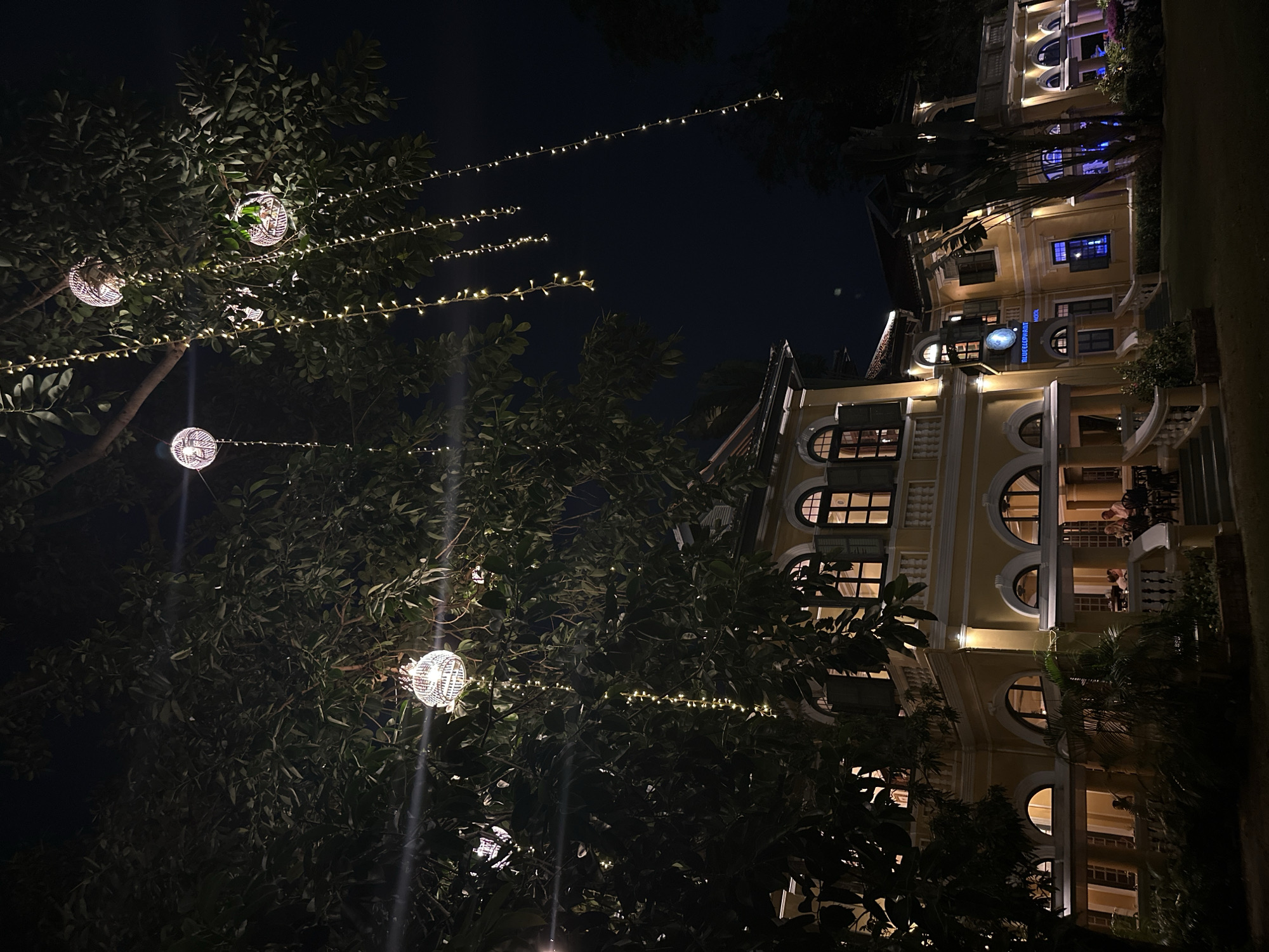 At night in Phuket City Municipality, Thailand, a cream-colored building with arched windows is illuminated by interior lights. The building, partly obscured by dark trees and foliage, has a vertical sign with the words "BLUEELEPHANT" vertically stacked and the word "COOKING SCHOOL" below. Strings of small lights are hung in the trees, interspersed with spherical, cage-like light fixtures. The sky is dark. A few people are visible on the balcony of the building.