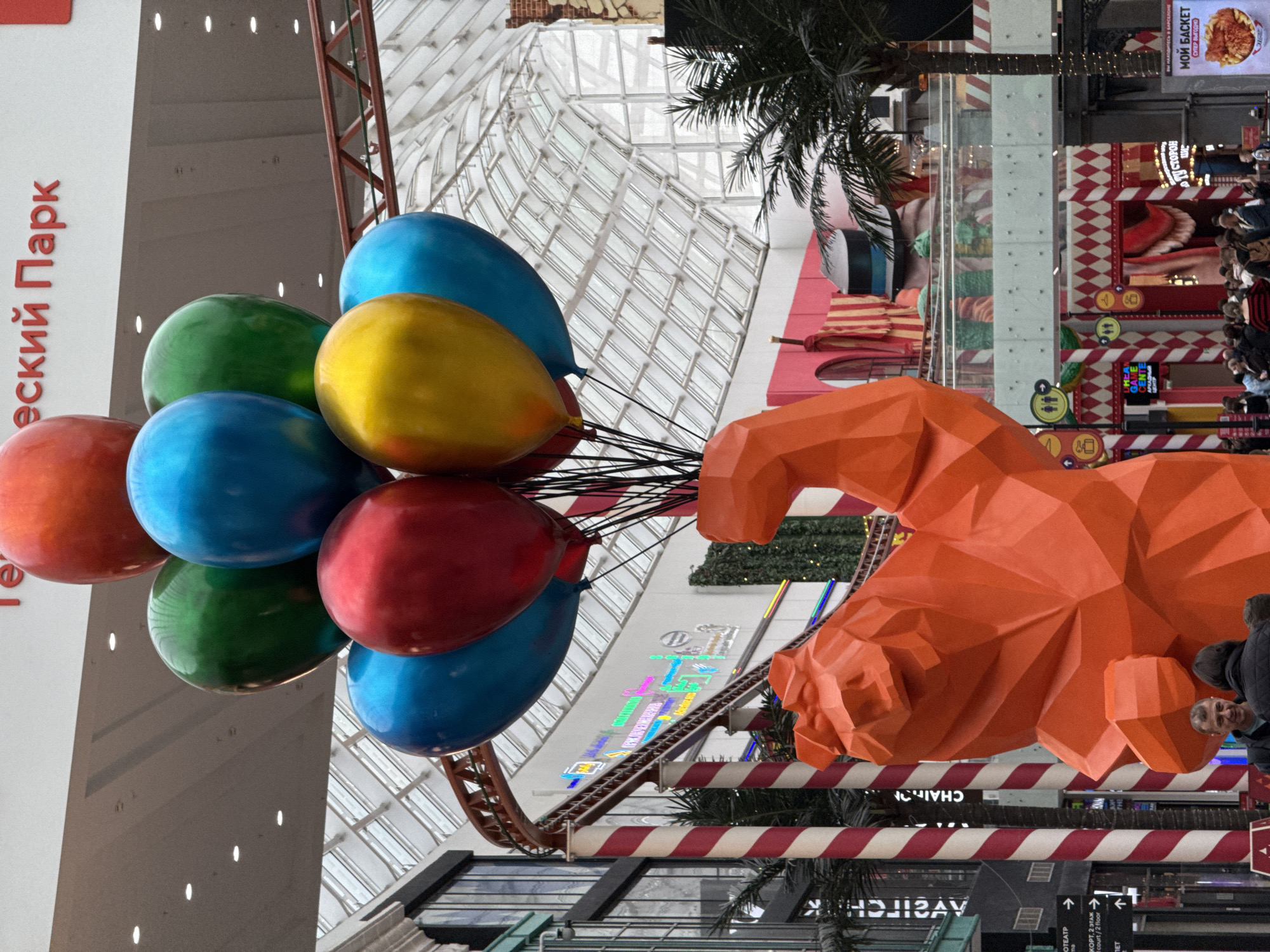 The image shows a view inside a large shopping mall, likely in Moscow, Russia, based on the Cyrillic text visible in the signage. There is a cluster of colorful balloons – red, blue, green, and yellow – positioned on the left side of the frame. An orange, angular sculpture of an animal head is prominent on the right side of the image.
The background reveals the mall's interior, featuring a complex network of escalators, walkways, retail spaces, and a glass ceiling allowing natural light to filter through. A rollercoaster track is visible in the midground, adding an amusement park element to the shopping environment. There is also a palm tree. There are people. There are also red and white striped decorations.